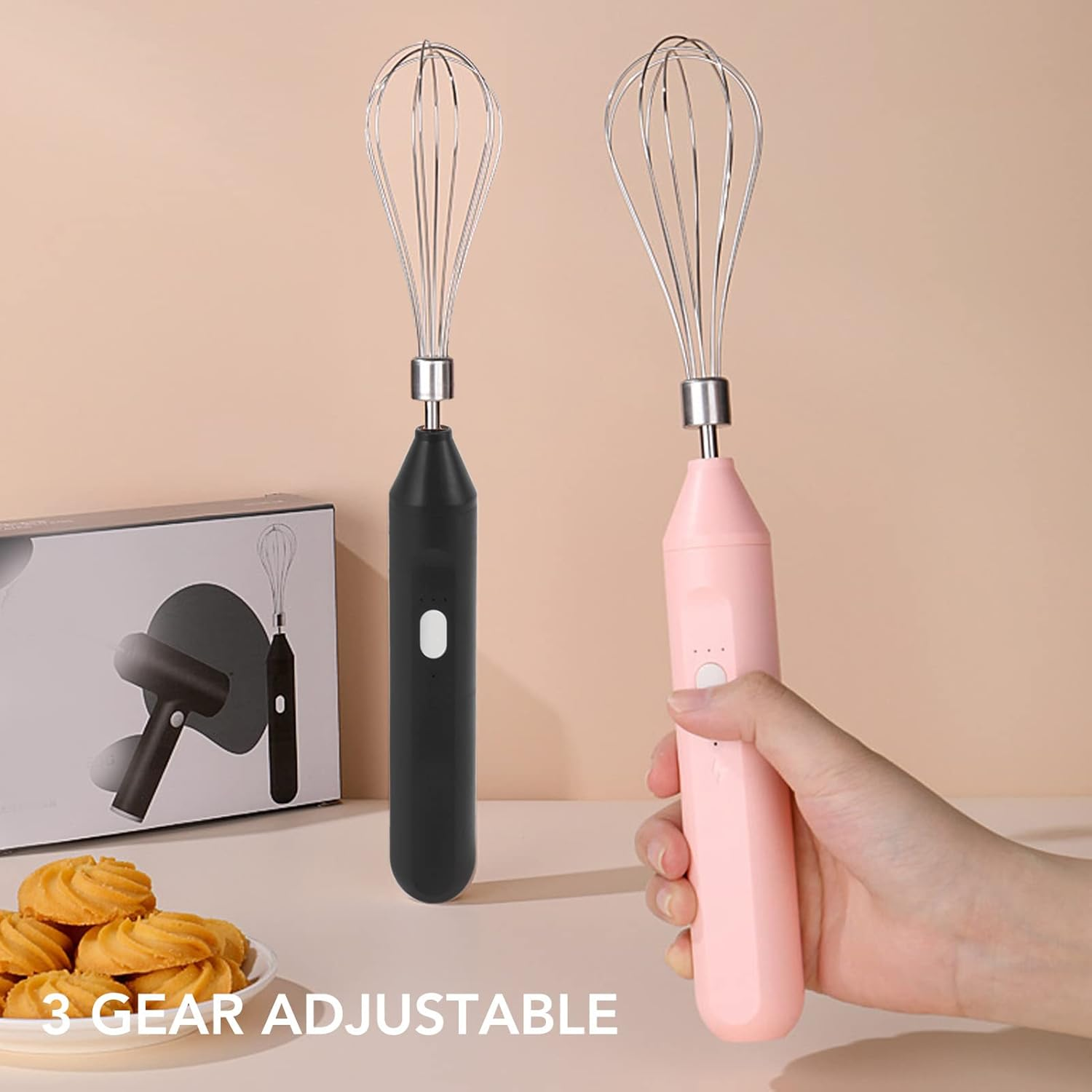 Zwei elektrische Schneebesen, einer schwarz und einer rosa, mit einer Schachtel und Keksen im Vordergrund. Text: "3 Gear Adjustable".