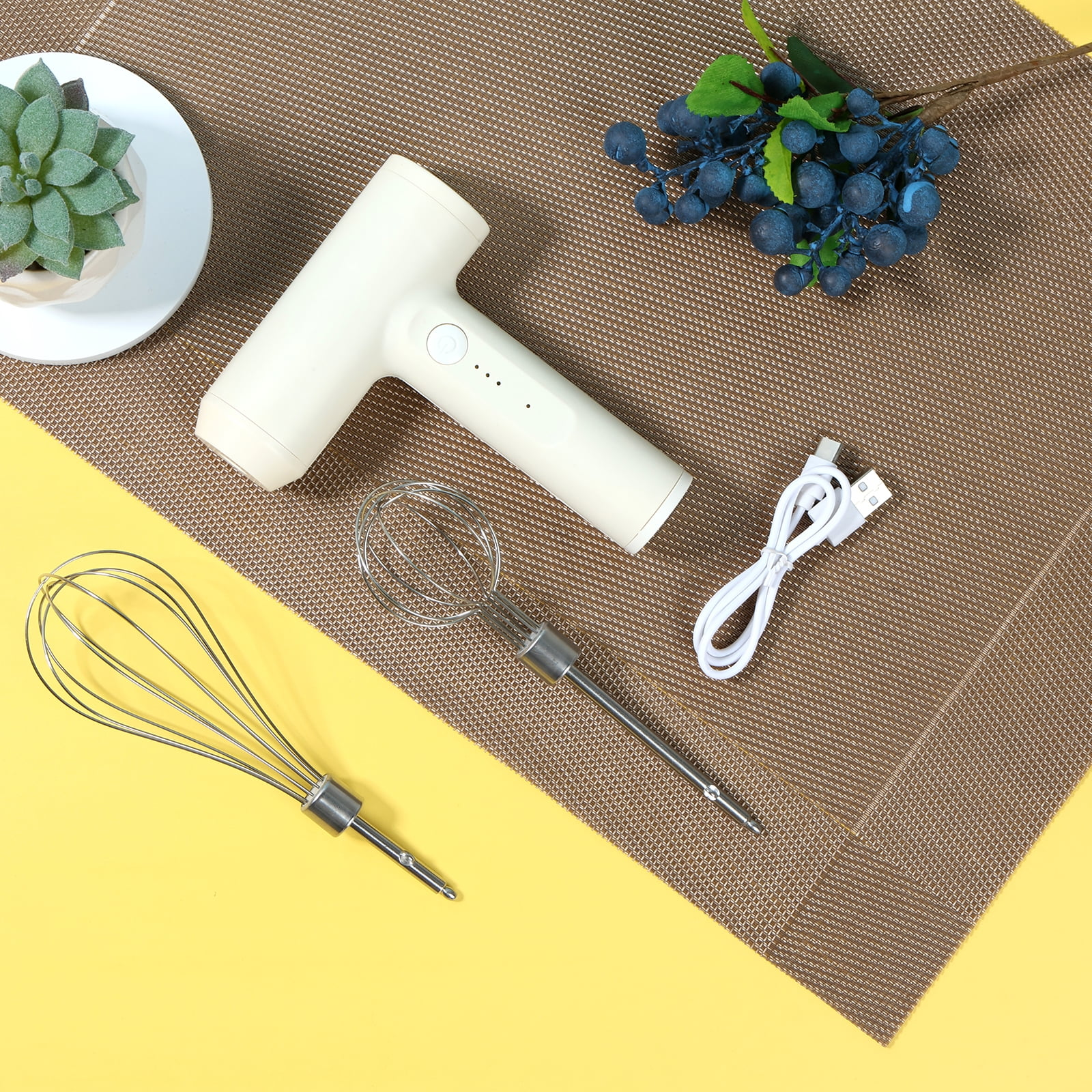 Ein Handmixer, Schneebesen und ein Ladekabel auf einer braunen Matte und gelben Oberfläche. Eine Pflanze und Beeren sind ebenfalls vorhanden.