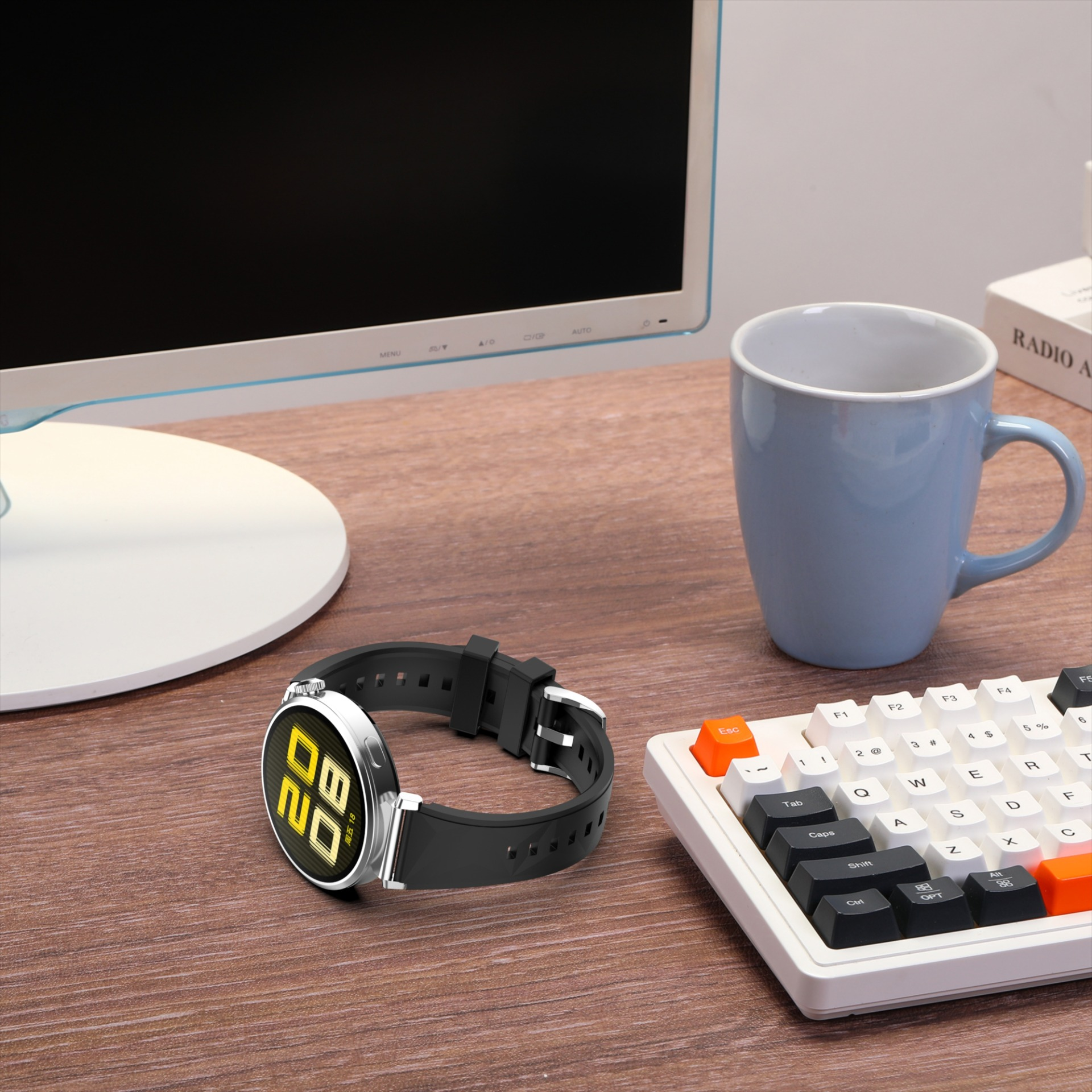 Uno smartwatch con cinturino nero è appoggiato su una scrivania di legno. Sul tavolo ci sono anche un monitor, una tazza e una tastiera.
