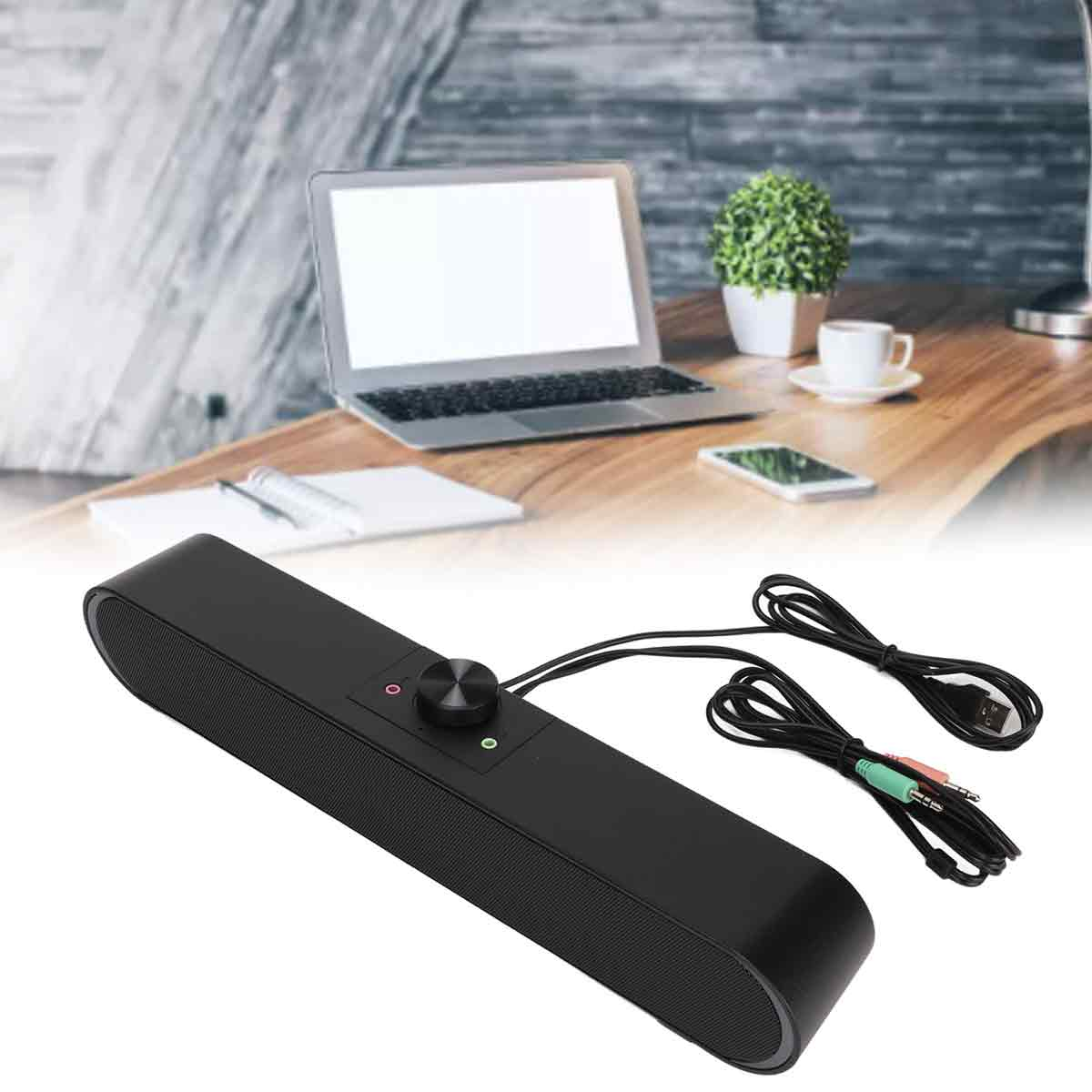 Ein schwarzer Lautsprecher mit Lautstärkeregler und Audiokabeln steht auf einem Holztisch. Ein Laptop, ein Telefon und eine kleine Pflanze sind sichtbar.