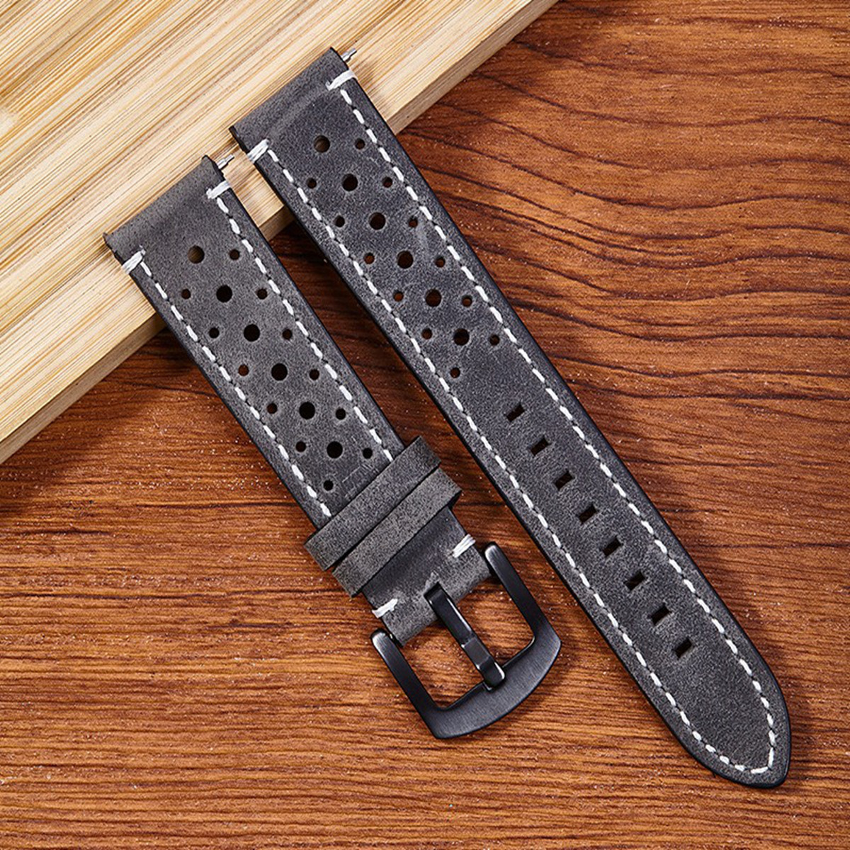 Due cinturini in pelle grigia con cuciture bianche e fori su una superficie di legno. Uno ha una fibbia nera.