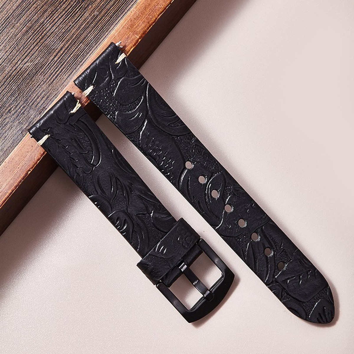 Cinturino per orologio in pelle nera con motivo floreale, fibbia e cuciture. Su superficie rosa chiaro, vicino a cornice in legno.