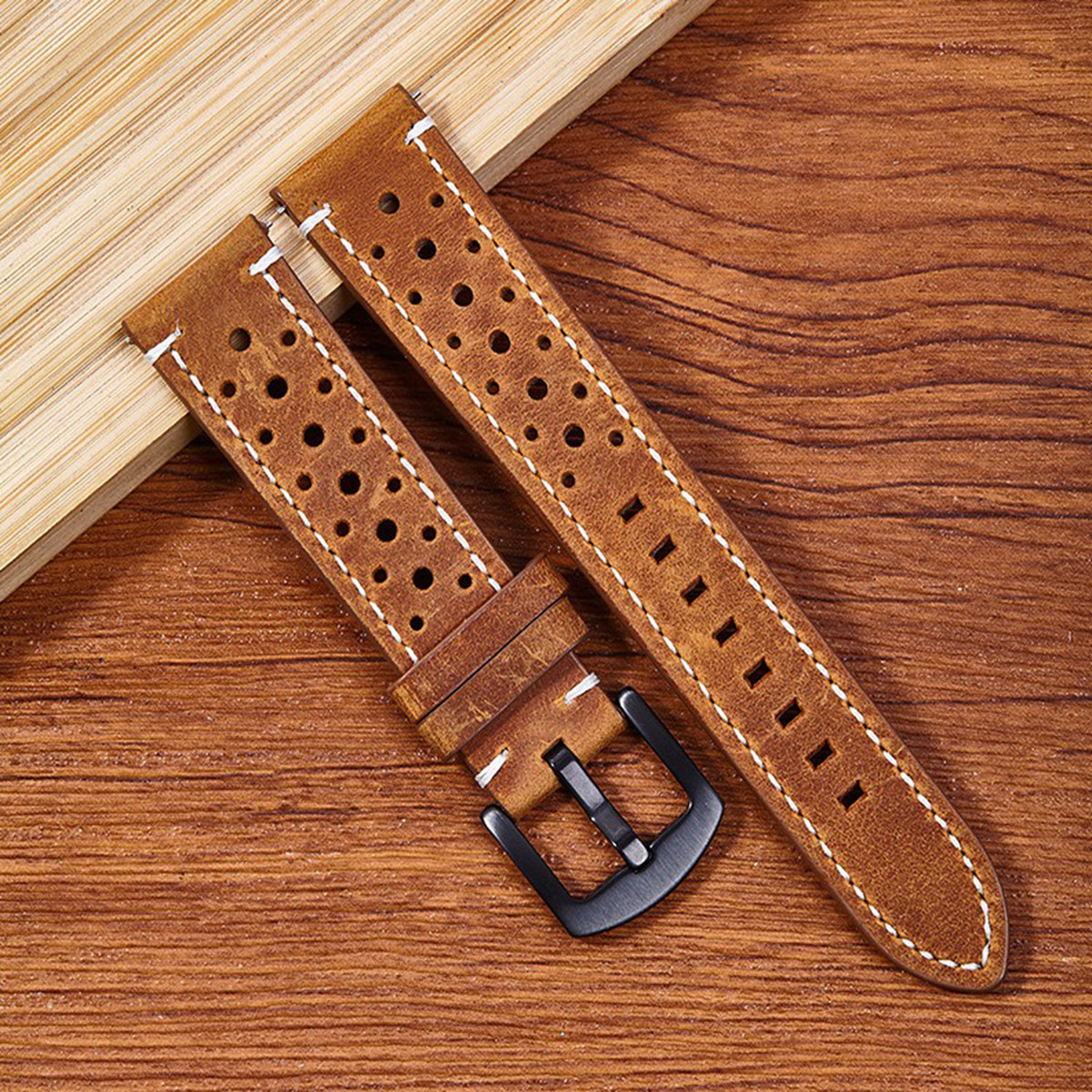 Cinturini per orologi in pelle marrone con cuciture bianche e fibbia nera, su superficie in legno. Design perforato.
