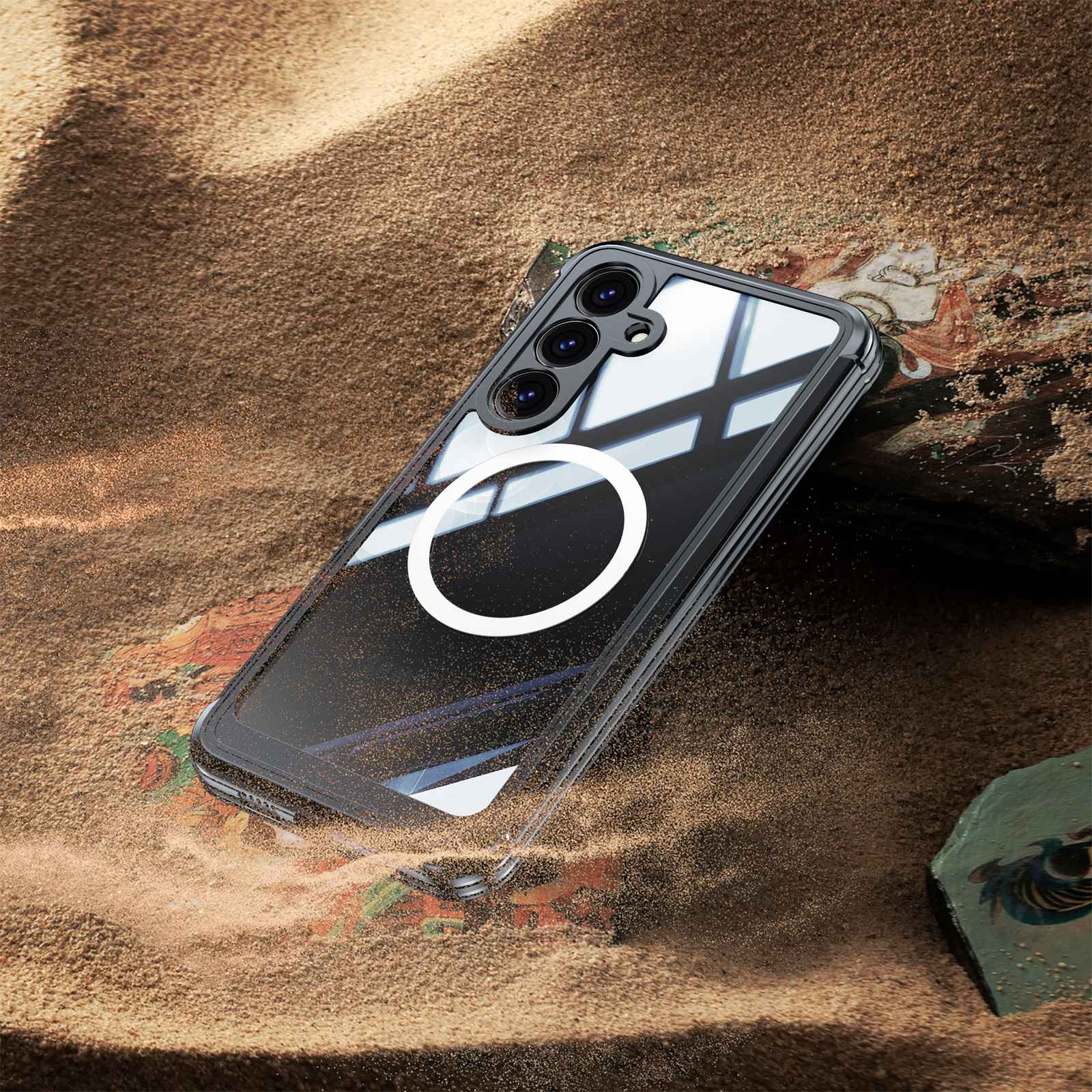 Ein Smartphone in einer durchsichtigen Hülle liegt auf Sand. Die Hülle hat einen weißen Ring. Der Hintergrund zeigt Sand und einen Stein.