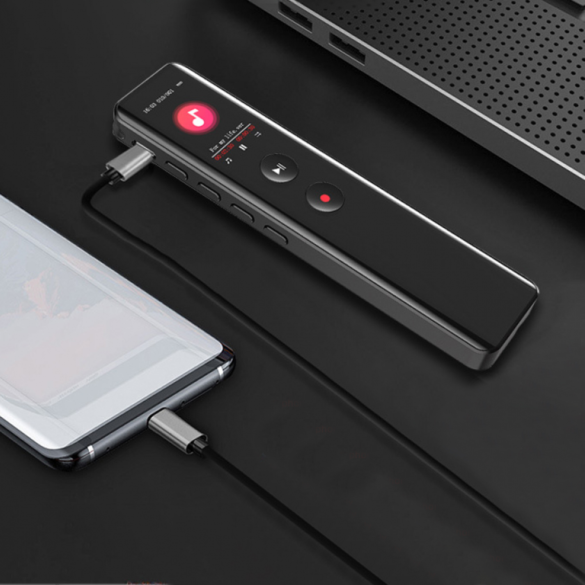 Ein schwarzer Audioplayer, der über ein Kabel mit einem Telefon verbunden ist. Im Hintergrund befindet sich ein Laptop.