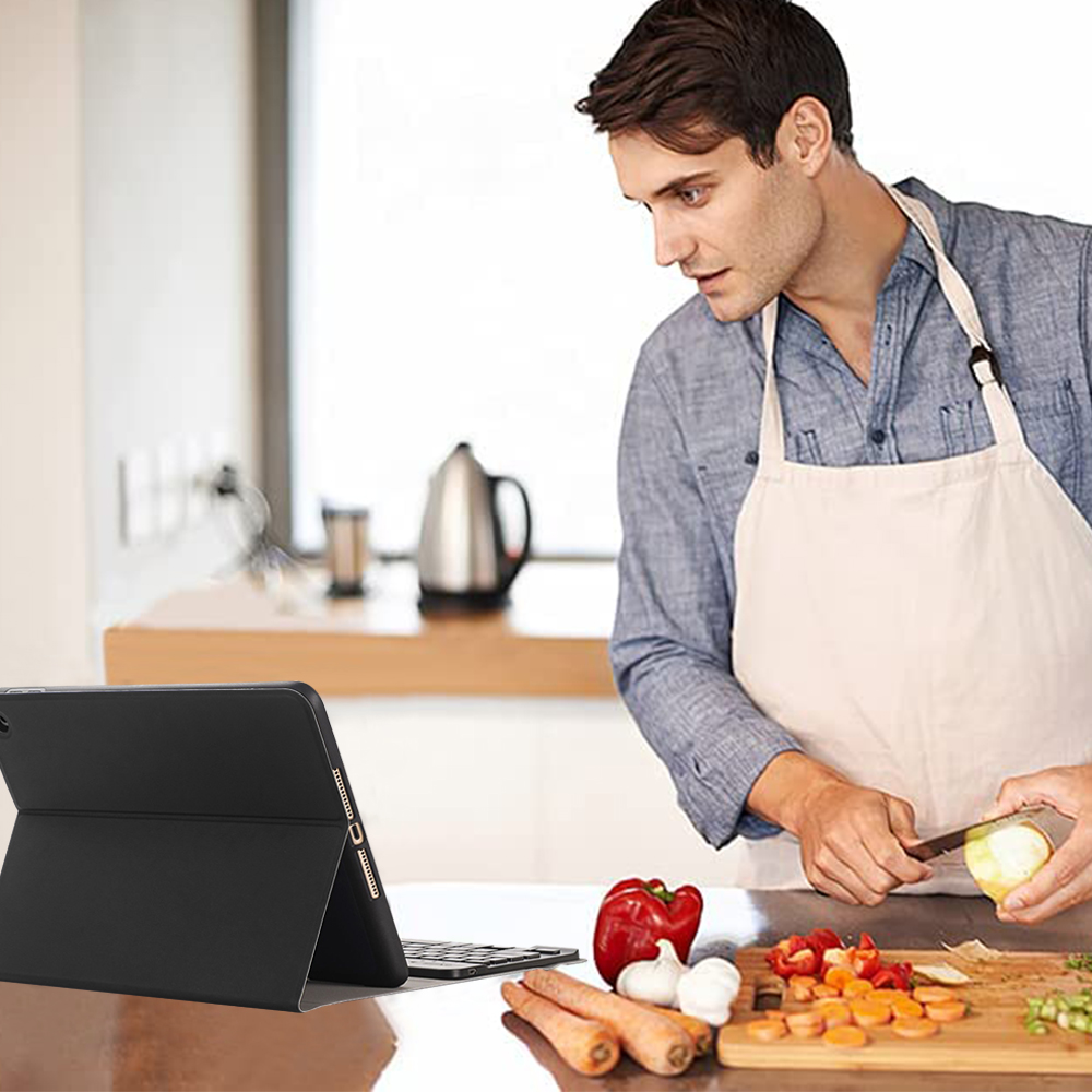 Mann mit Schürze schält Obst, mit Gemüse und einem Tablet im Vordergrund. Küchenhintergrund mit einem Wasserkocher.