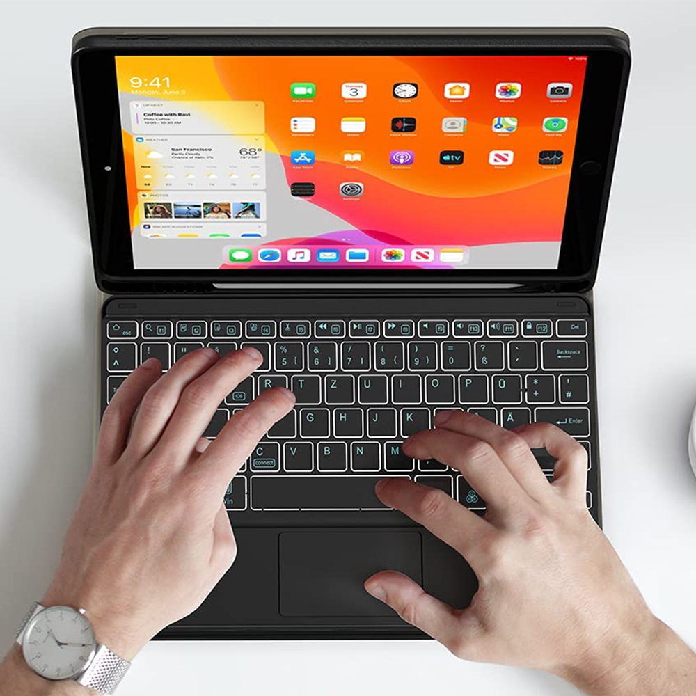 Hände tippen auf einer beleuchteten Tastatur, die an ein iPad angeschlossen ist und den Startbildschirm zeigt. Eine Uhr ist am Handgelenk zu sehen. Weißer Hintergrund.