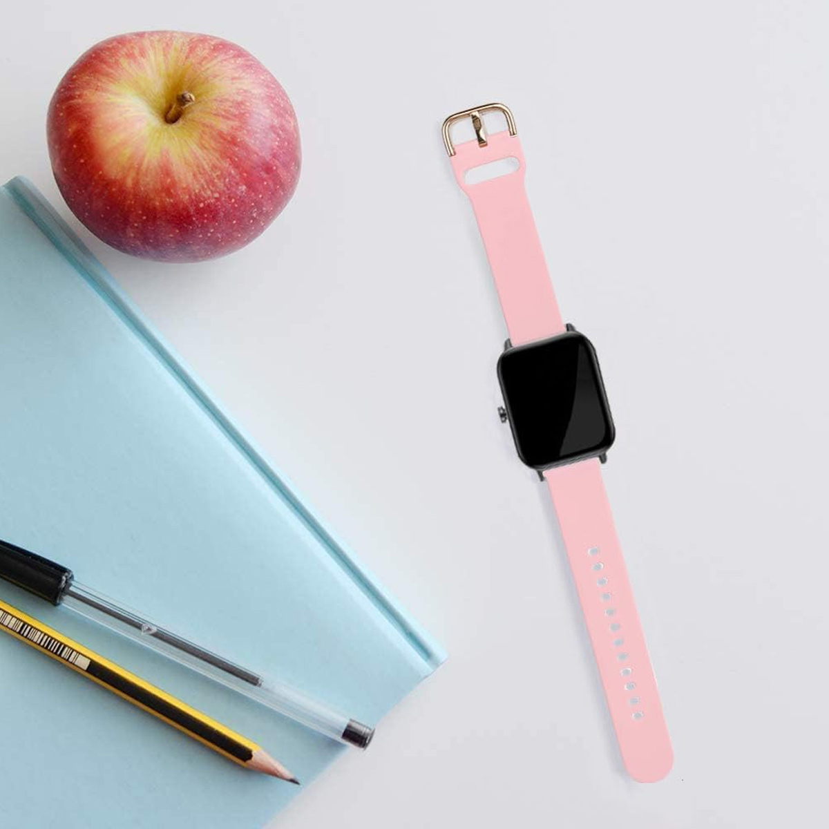 Ein Apfel, eine rosa Uhr, ein blaues Notizbuch, ein Stift und ein Bleistift liegen auf einer weißen Oberfläche.