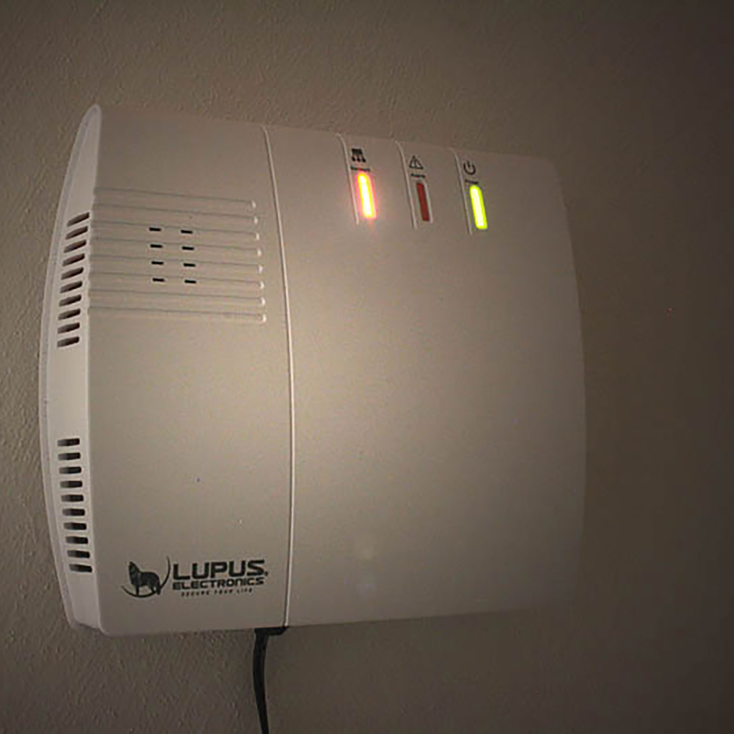 Ein weißes Lupus Electronics-Gerät ist an einer Wand montiert. Es hat Kontrollleuchten und ein Logo. Ein schwarzes Kabel geht davon ab.