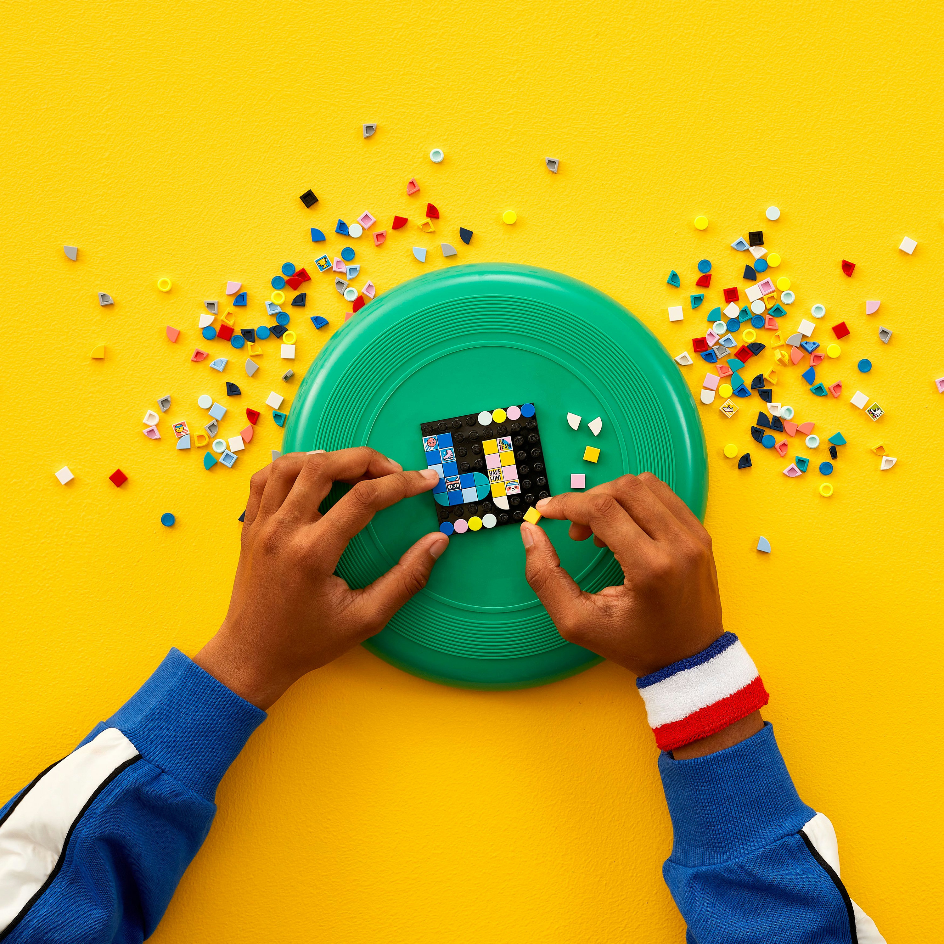 Hands assembling a Lego design on a green frisbee, surrounded by scattered colorful Lego pieces on a yellow background.