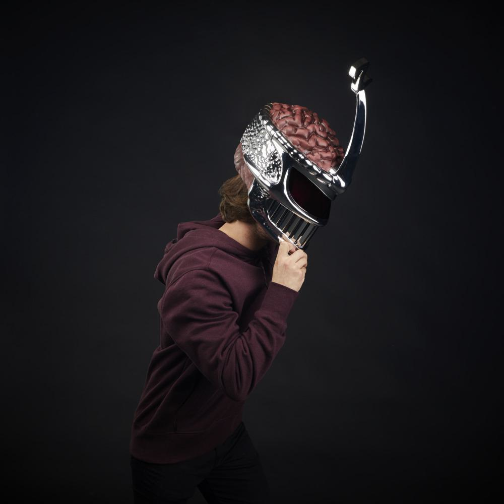 Burgundy kapüşonlu bir kişi, beyne benzeyen gümüş ve kırmızı bir kask tutuyor. Arka plan düz siyah.