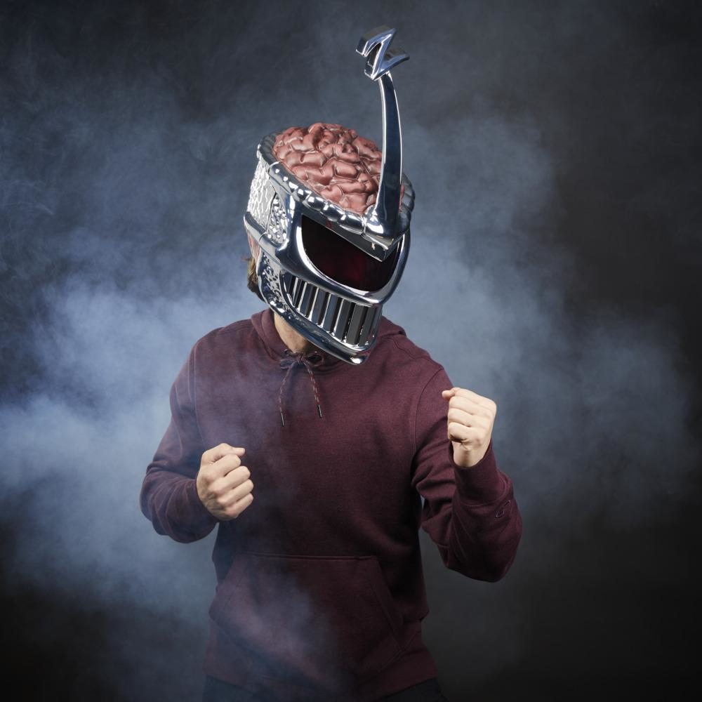 Üzerinde beyne benzer bir nesne bulunan gümüş ve kırmızı bir kask takan, bordo kapüşonlu bir sweatshirt giyen, yumrukları sıkılı, dumanlı bir arka planın önünde duran bir kişi.