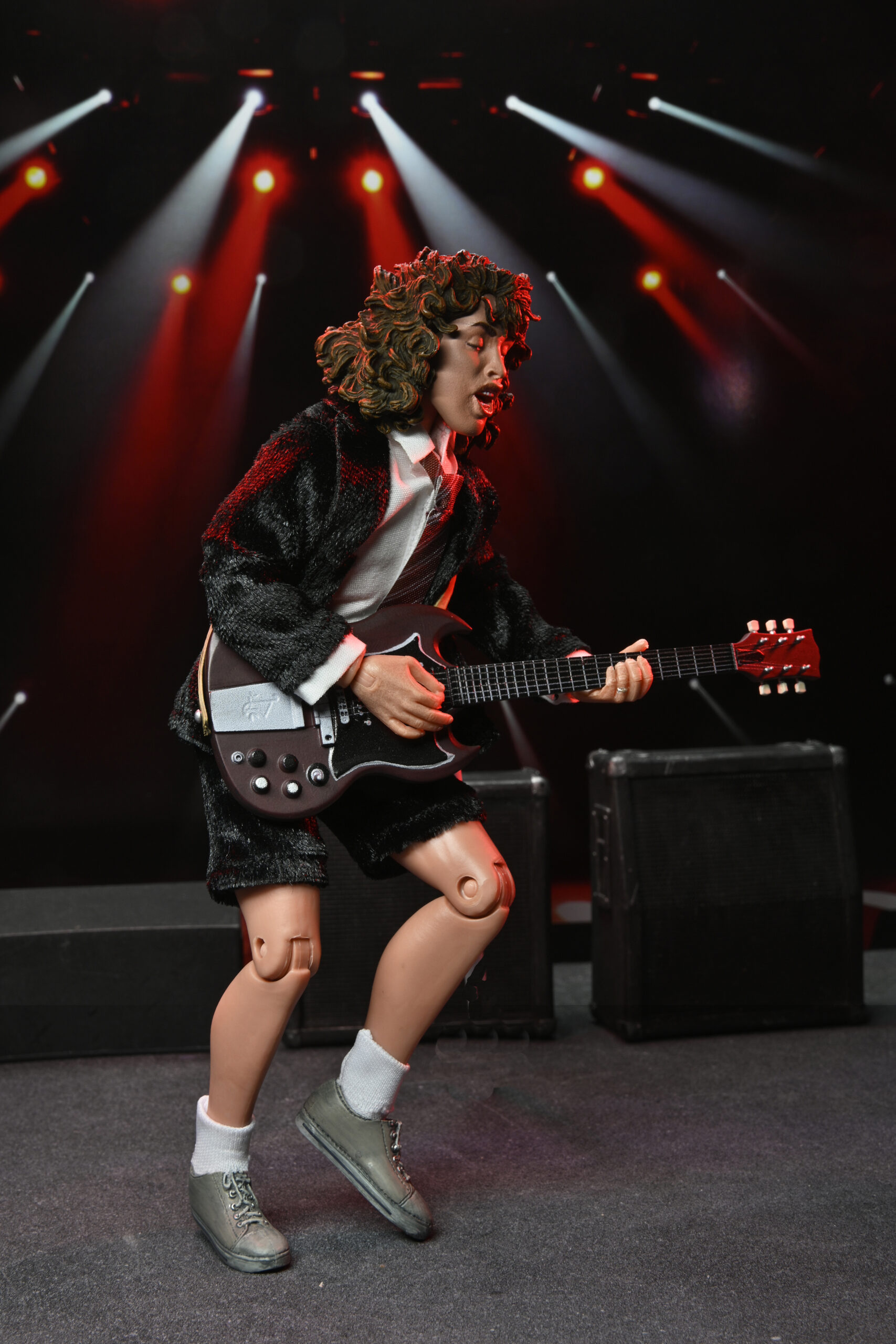 Bir figür sahnede gitar çalıyor. Siyah ceket, şort ve ayakkabı giyiyor. Yukarıdan kırmızı ışıklar parlıyor.