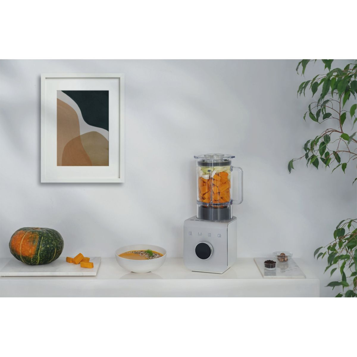 Escena de encimera de cocina. Una licuadora con trozos de calabaza. Calabaza, sopa y arte en la encimera. Planta verde al fondo.