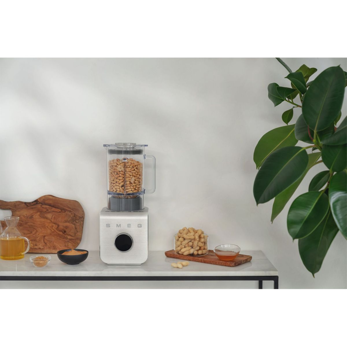 Escena de cocina con una licuadora llena de cacahuetes. Ingredientes y una planta en la encimera. Un tarro de aceite y un bol con mantequilla de cacahuete.