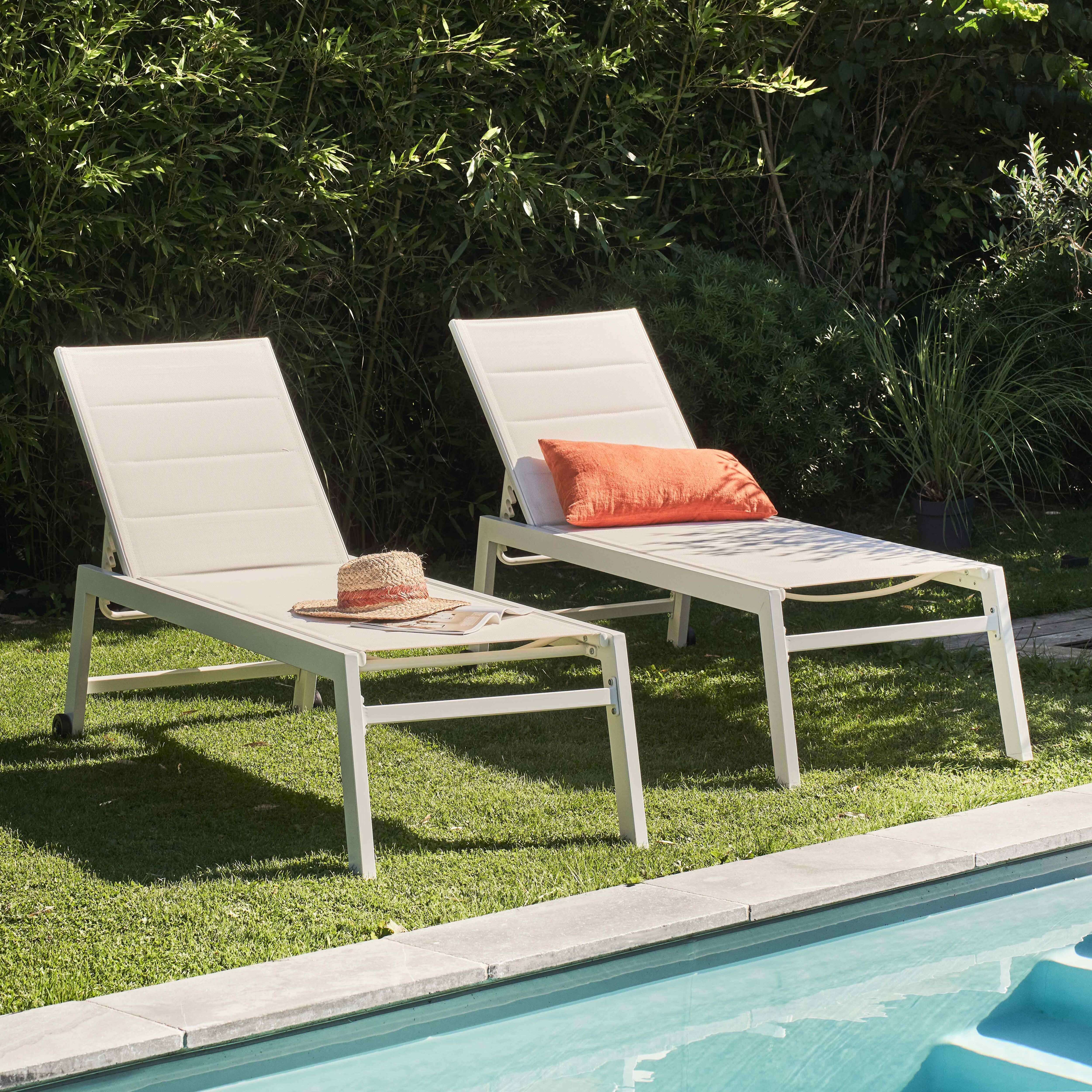 Zwei beige Liegestühle mit Strohhut und orangefarbenem Kissen auf grünem Gras neben einem Pool.