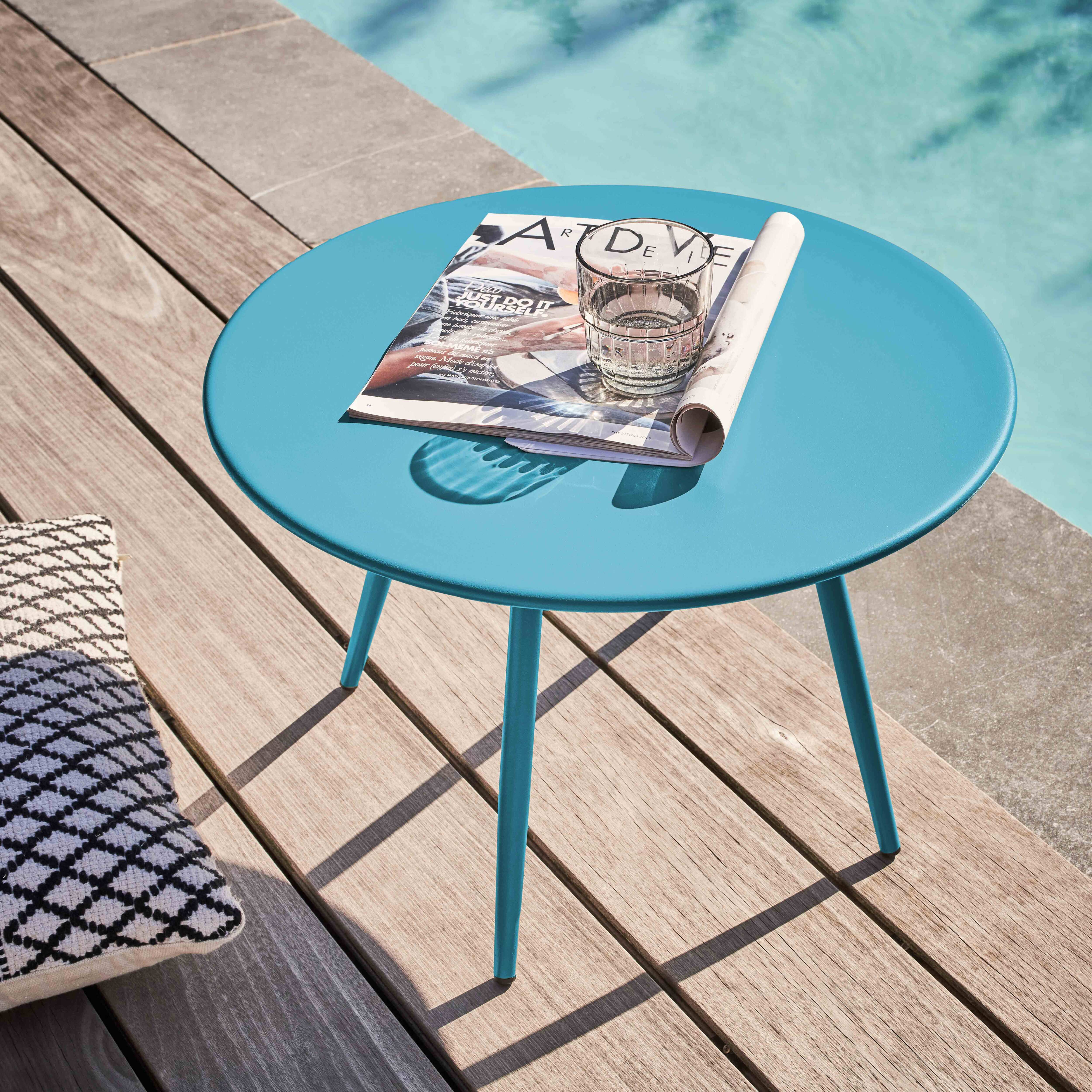 Ein blauer Tisch mit Zeitschrift und Glas steht auf Holz, neben einem Pool und einem gemusterten Kissen.