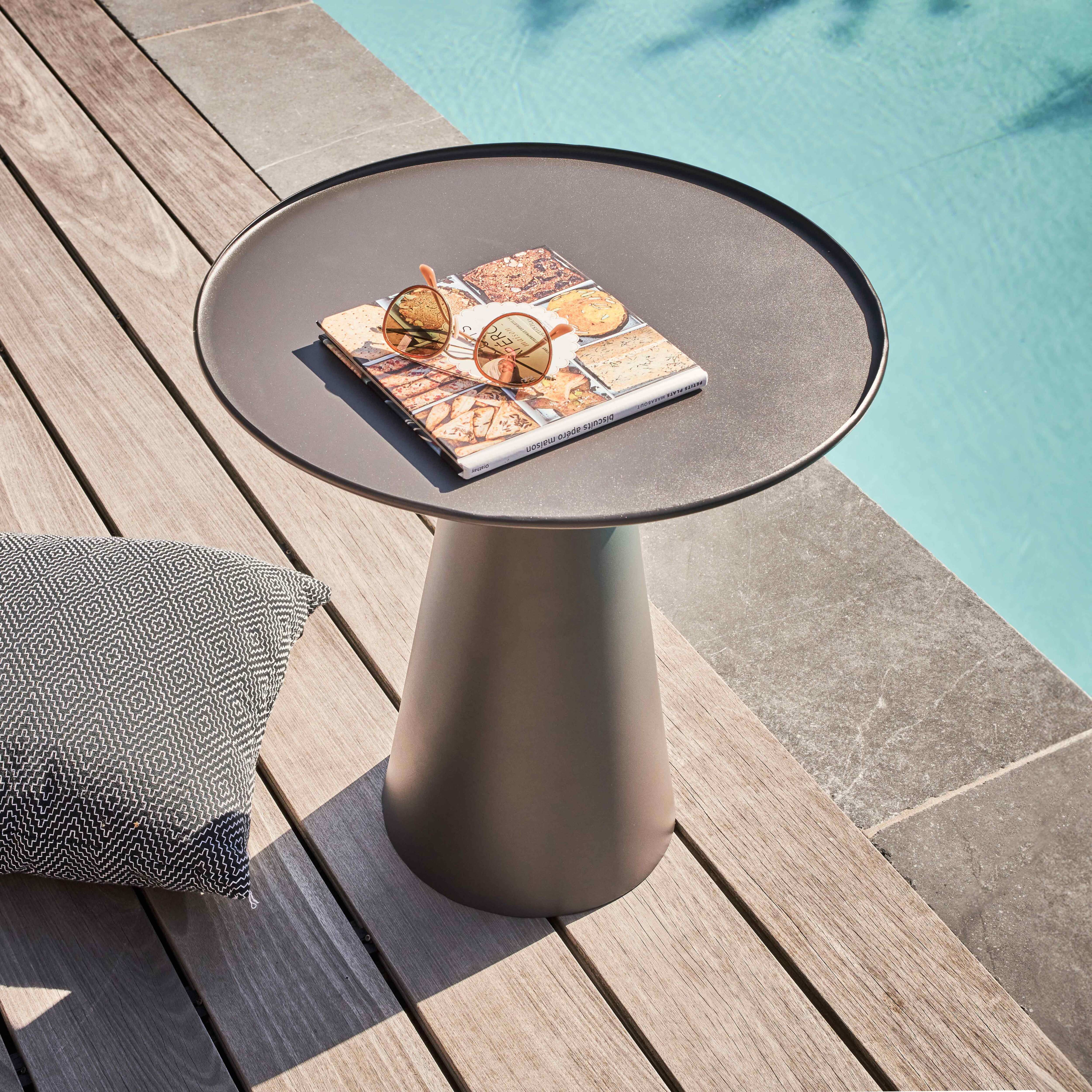 Ein kleiner grauer Tisch mit einer Zeitschrift und Sonnenbrille darauf. Ein Kissen liegt auf dem Holzdeck neben einem Pool.
