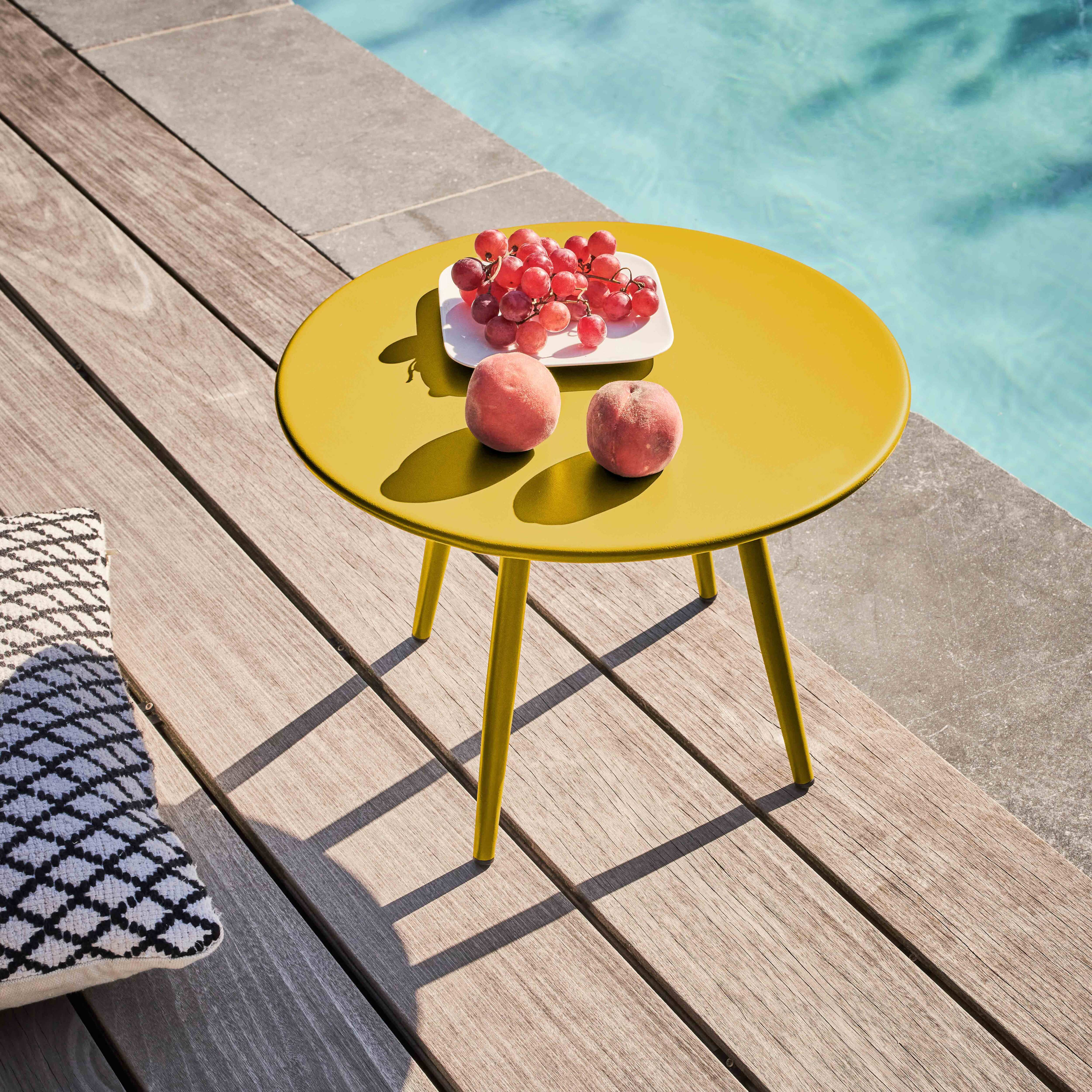 Ein gelber Tisch mit Trauben, Pfirsichen und einem Kissen auf Holzplanken neben einem Pool.