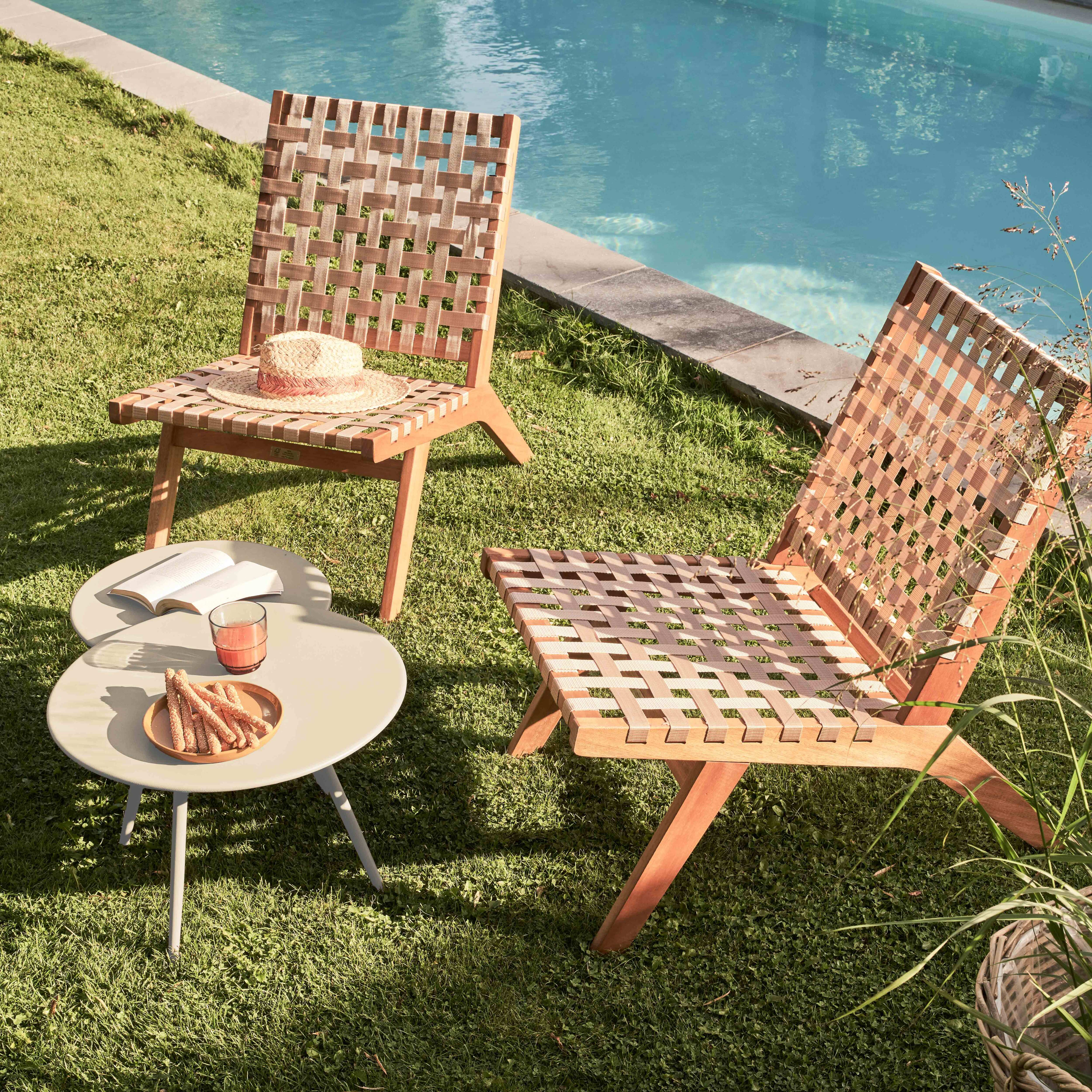 Zwei Stühle mit gewebten Sitzen, ein Tisch mit Snacks und Getränken und ein Pool im Hintergrund, alles auf einer Wiese.