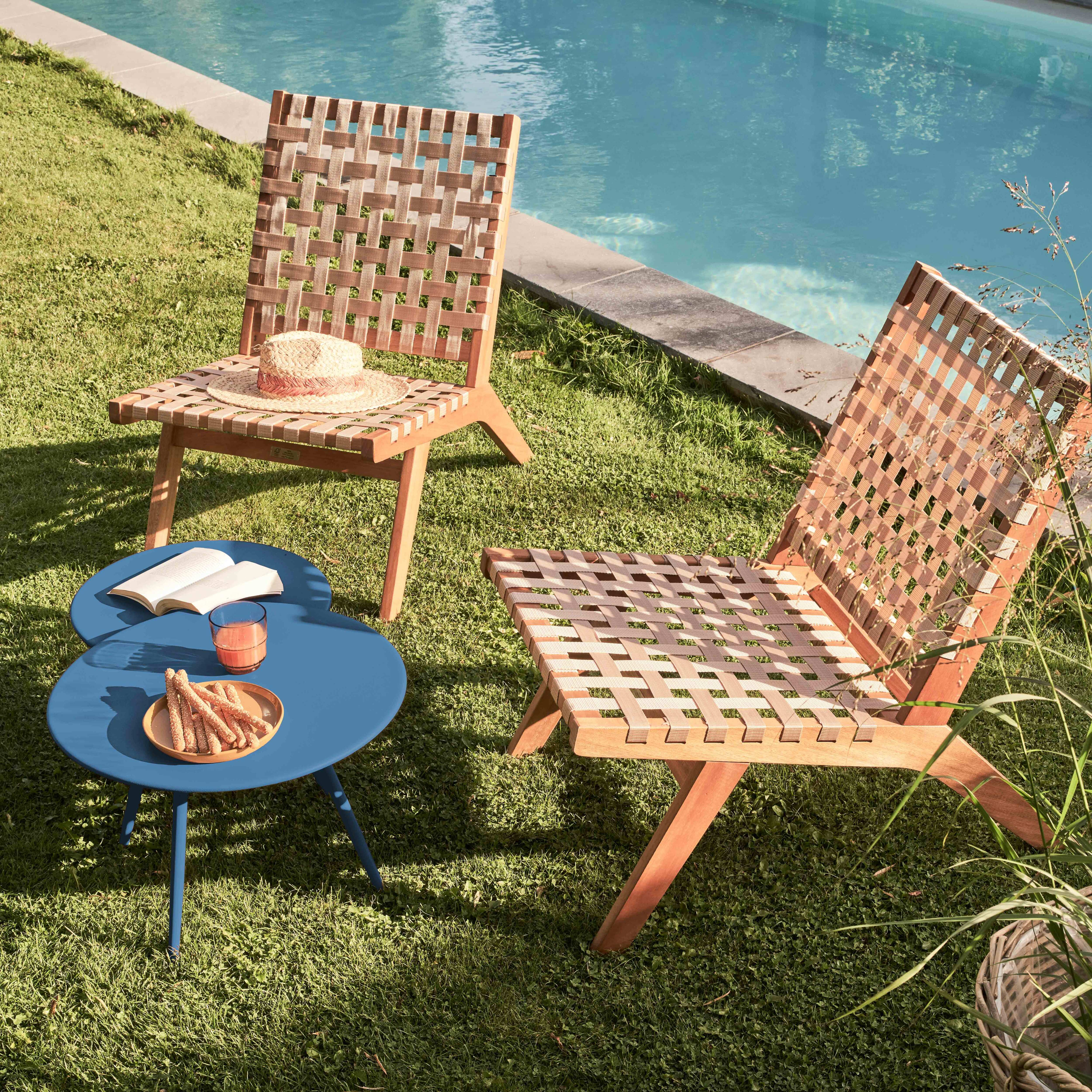 Szene im Freien mit gewebten Stühlen, einem blauen Tisch und einem Pool im Hintergrund. Ein Hut und Snacks liegen auf dem Tisch.