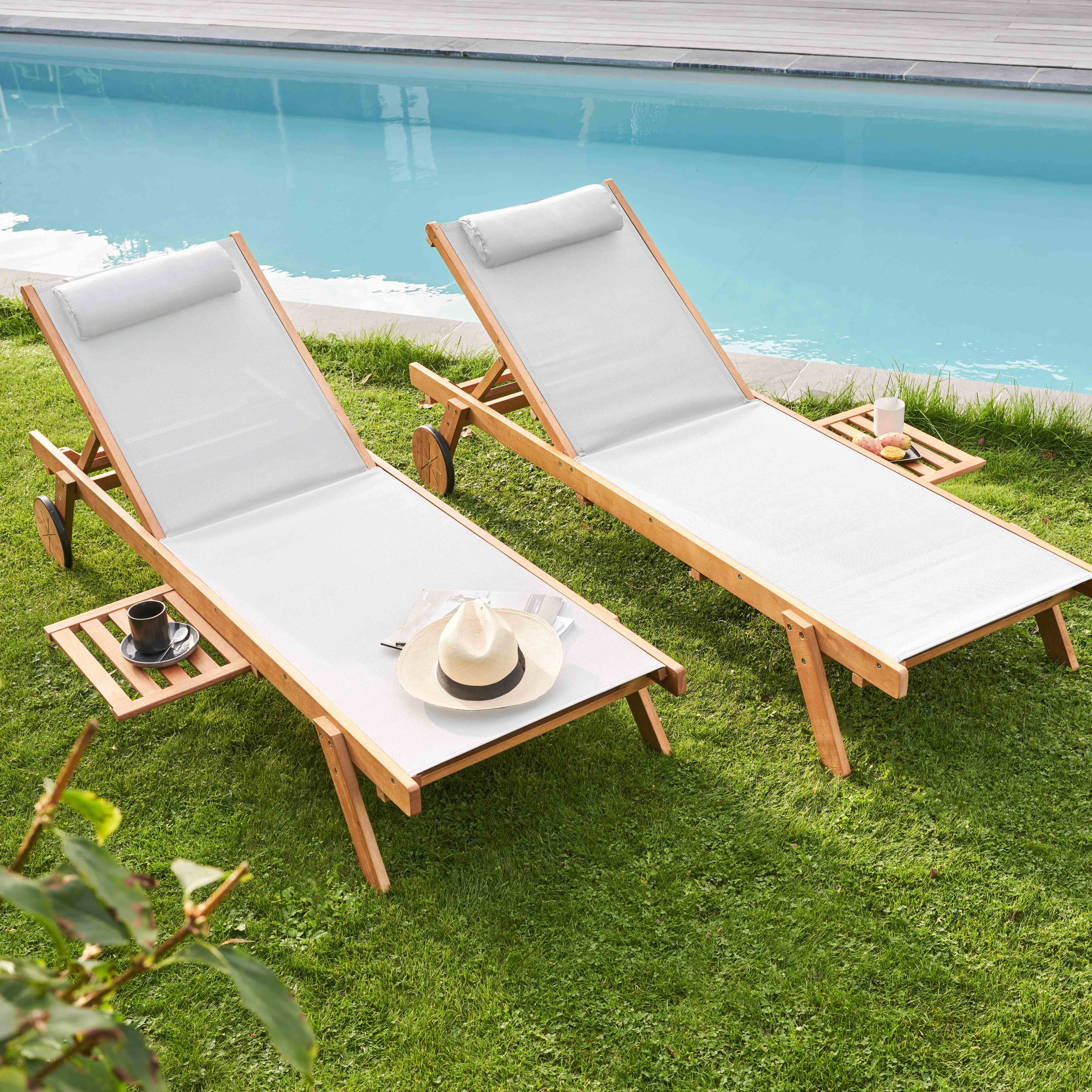 Zwei Holzliegen auf grünem Gras in der Nähe eines Pools. Ein Hut und eine Kaffeetasse liegen auf den Beistelltischen.