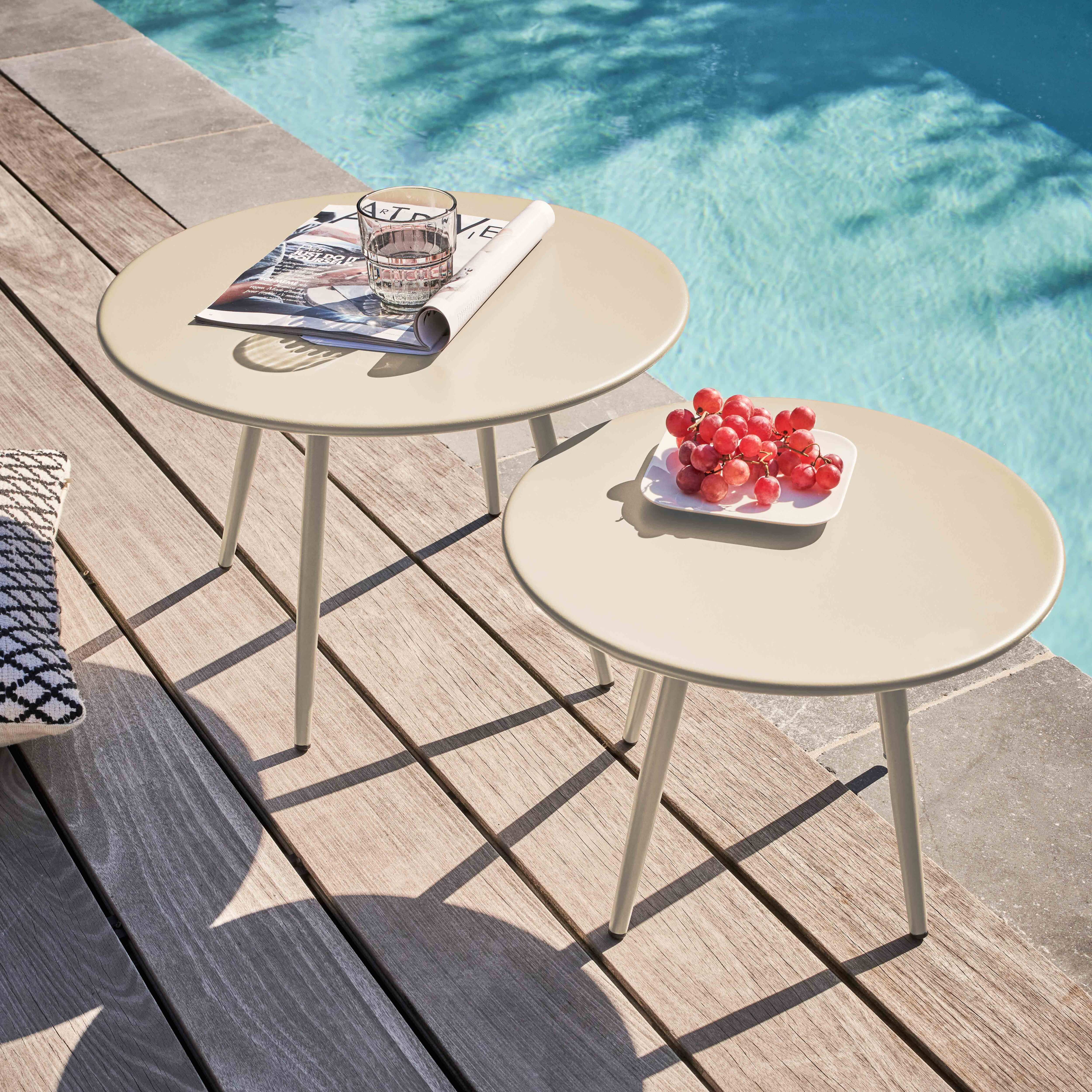 Zwei beige Tische mit Gegenständen auf einem Holzdeck am Pool. Einer hat ein Glas und eine Zeitschrift, der andere Trauben.