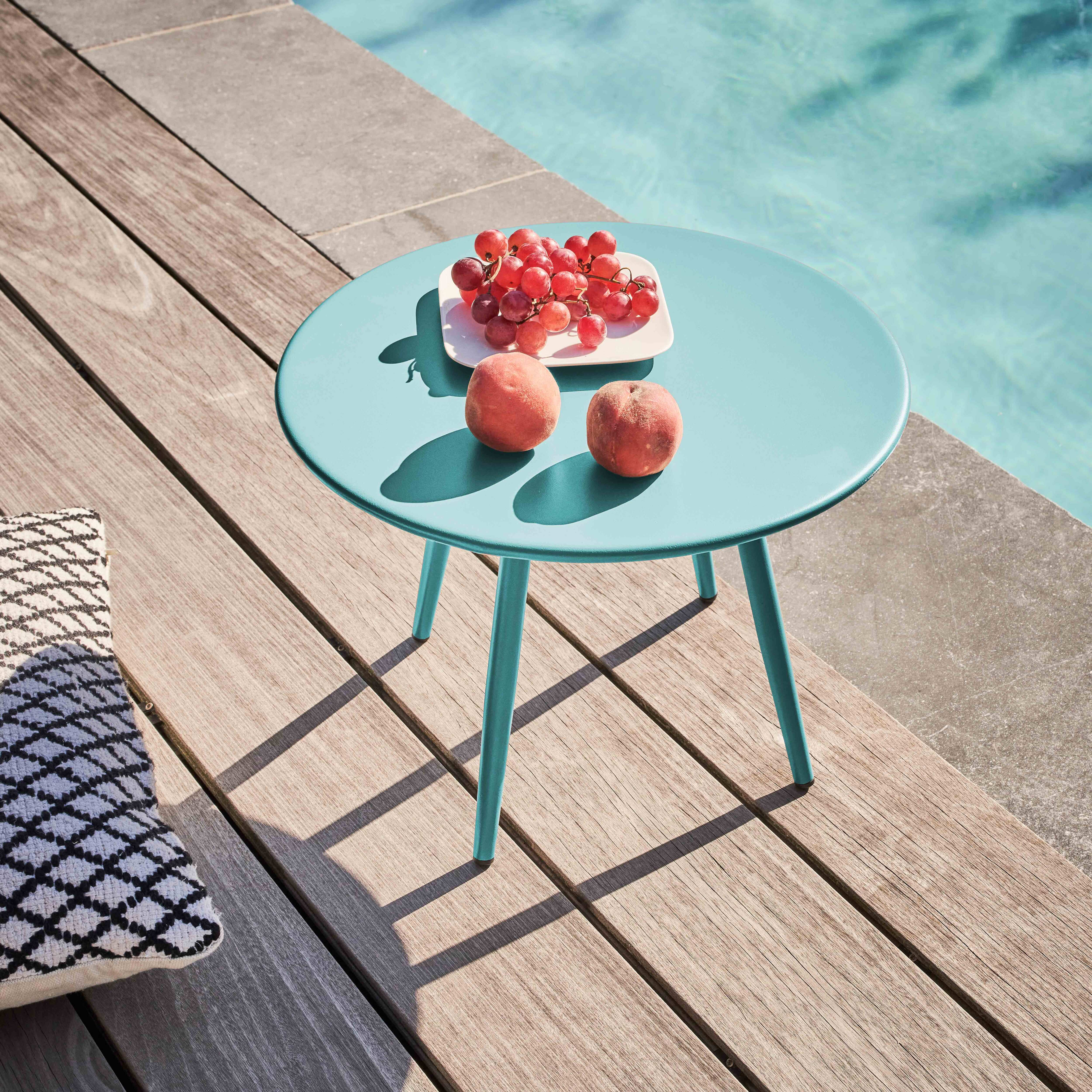 Ein türkisfarbener Tisch mit Trauben und Pfirsichen steht auf Holzplanken am Pool. Links ist ein gemustertes Kissen.
