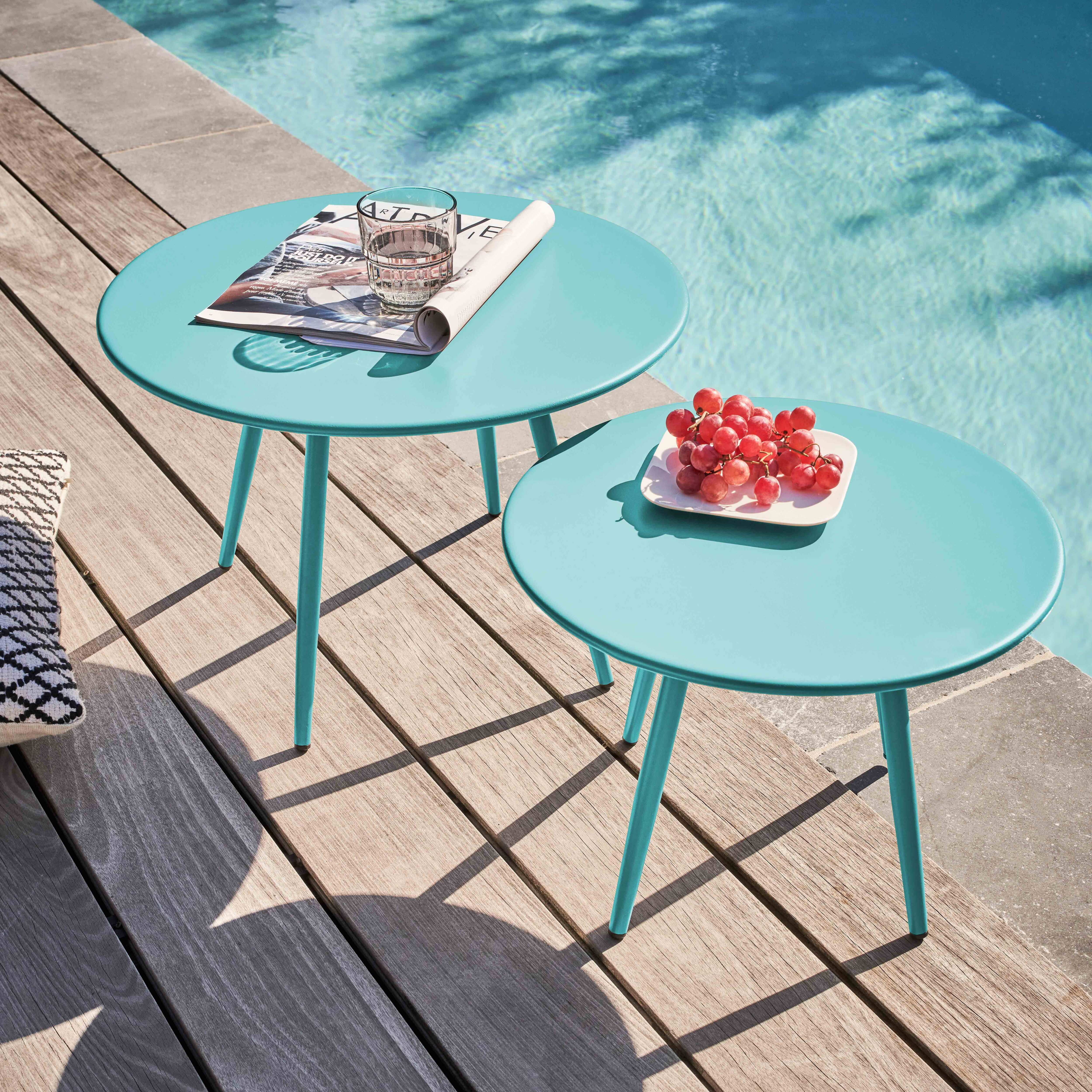 Zwei türkisfarbene Tische auf einem Holzdeck, einer mit Zeitschrift und Glas, der andere mit Trauben. Pool im Hintergrund.