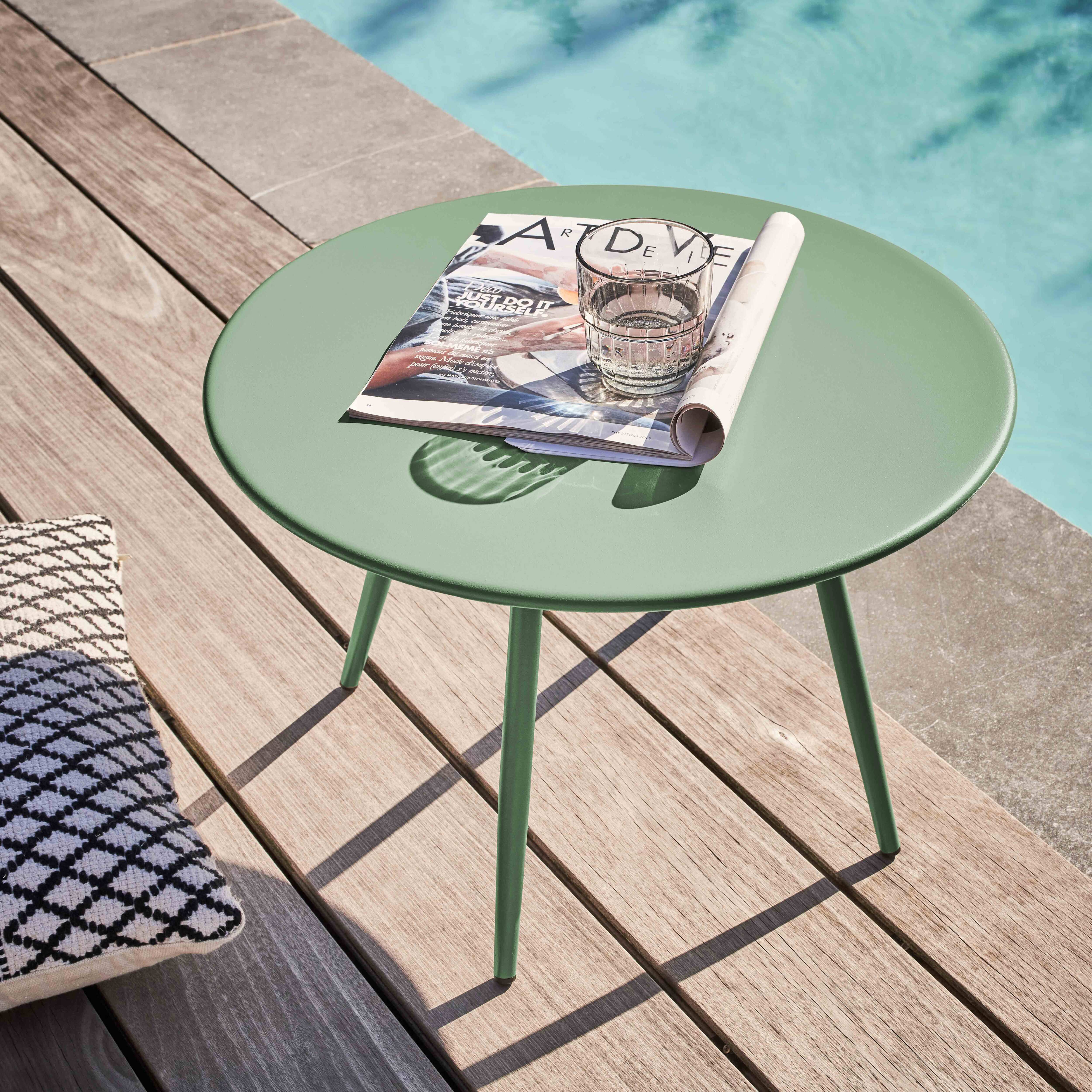 Ein grüner Tisch mit Magazin und Glas. Holzterrasse und Pool im Hintergrund. Ein Kissen liegt daneben.