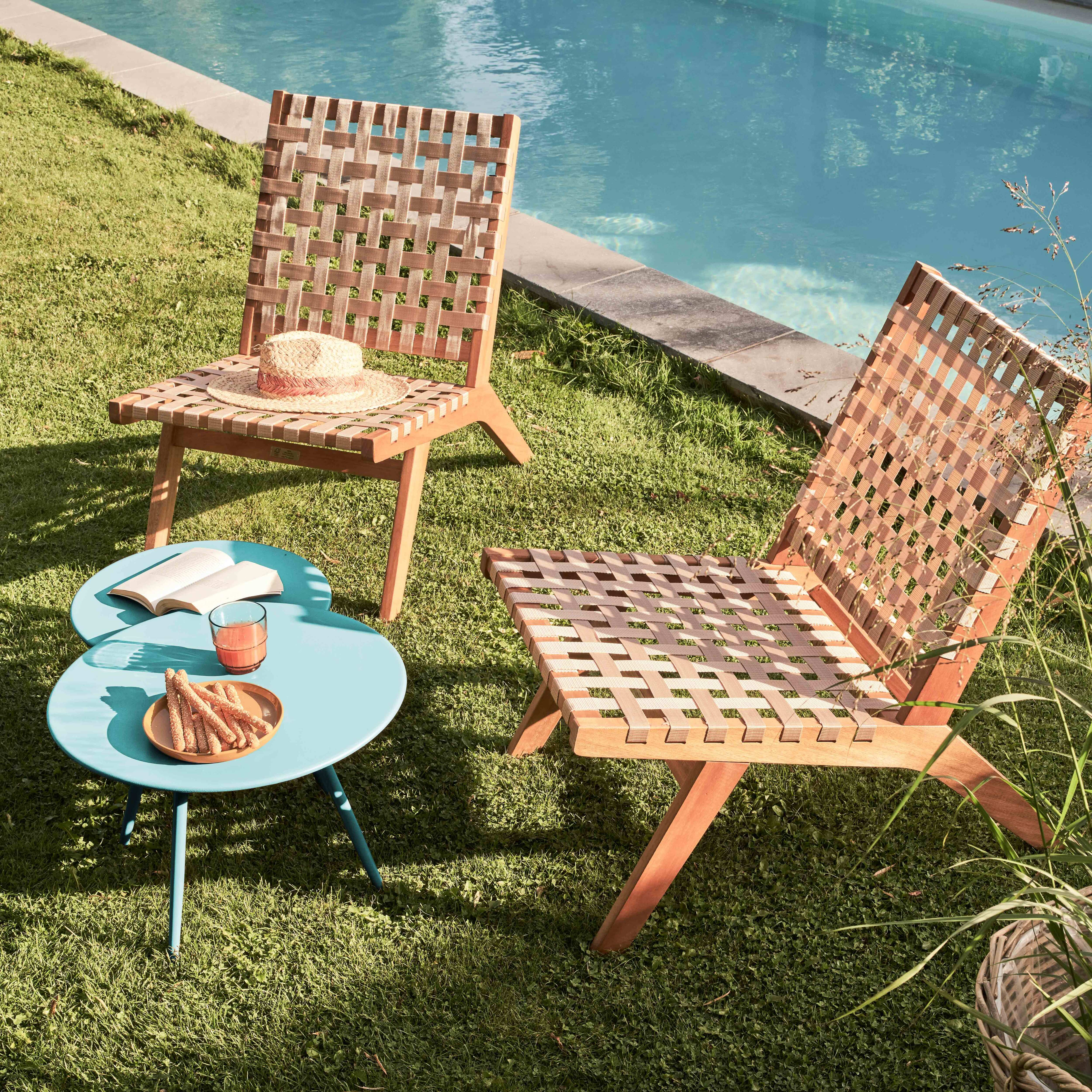 Zwei Holzstühle, ein blauer Tisch mit Snacks und einem Getränk, ein Swimmingpool und grünes Gras sind zu sehen.