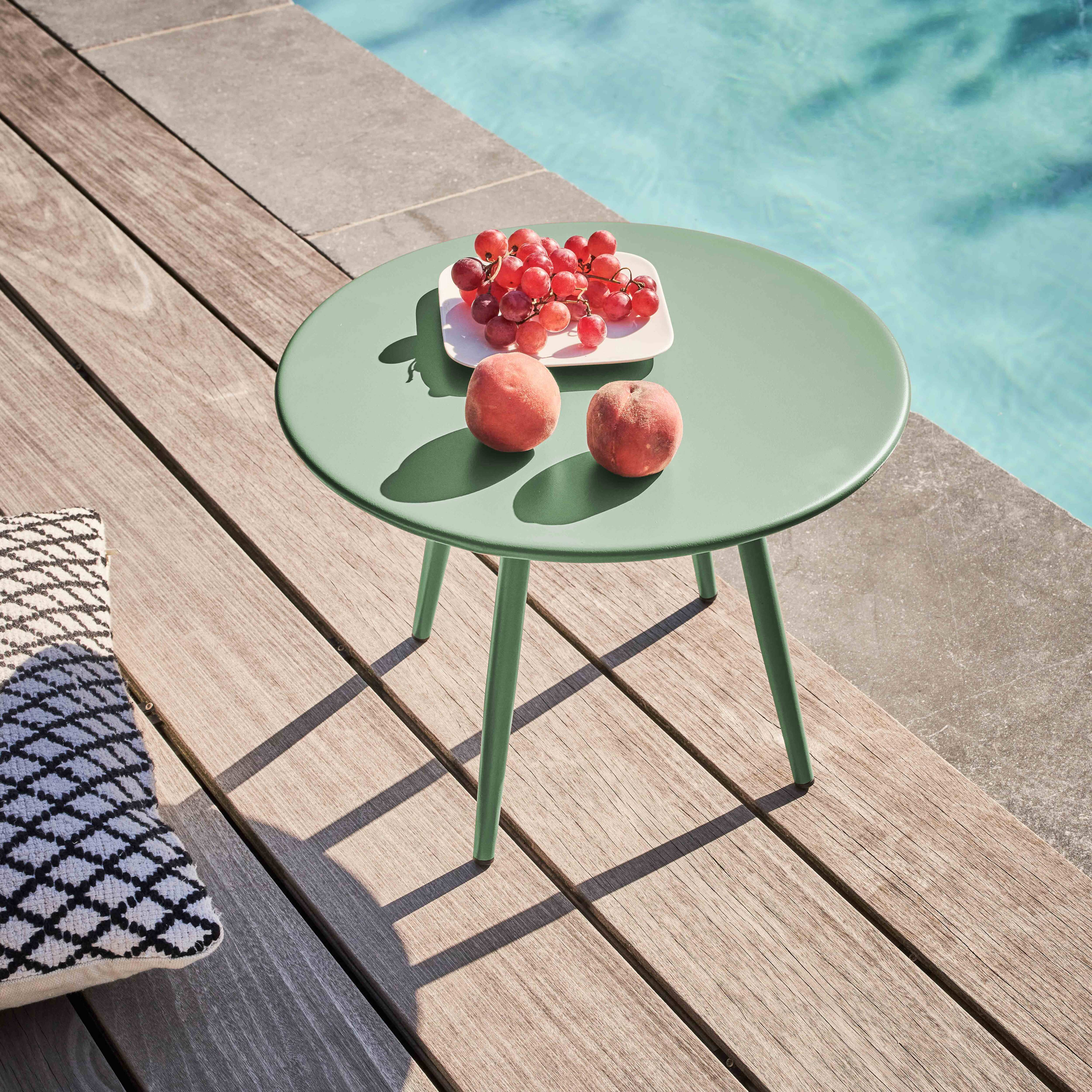 Ein grüner Tisch mit Trauben und Pfirsichen steht auf Holzplanken am Pool. Ein gemustertes Kissen links.