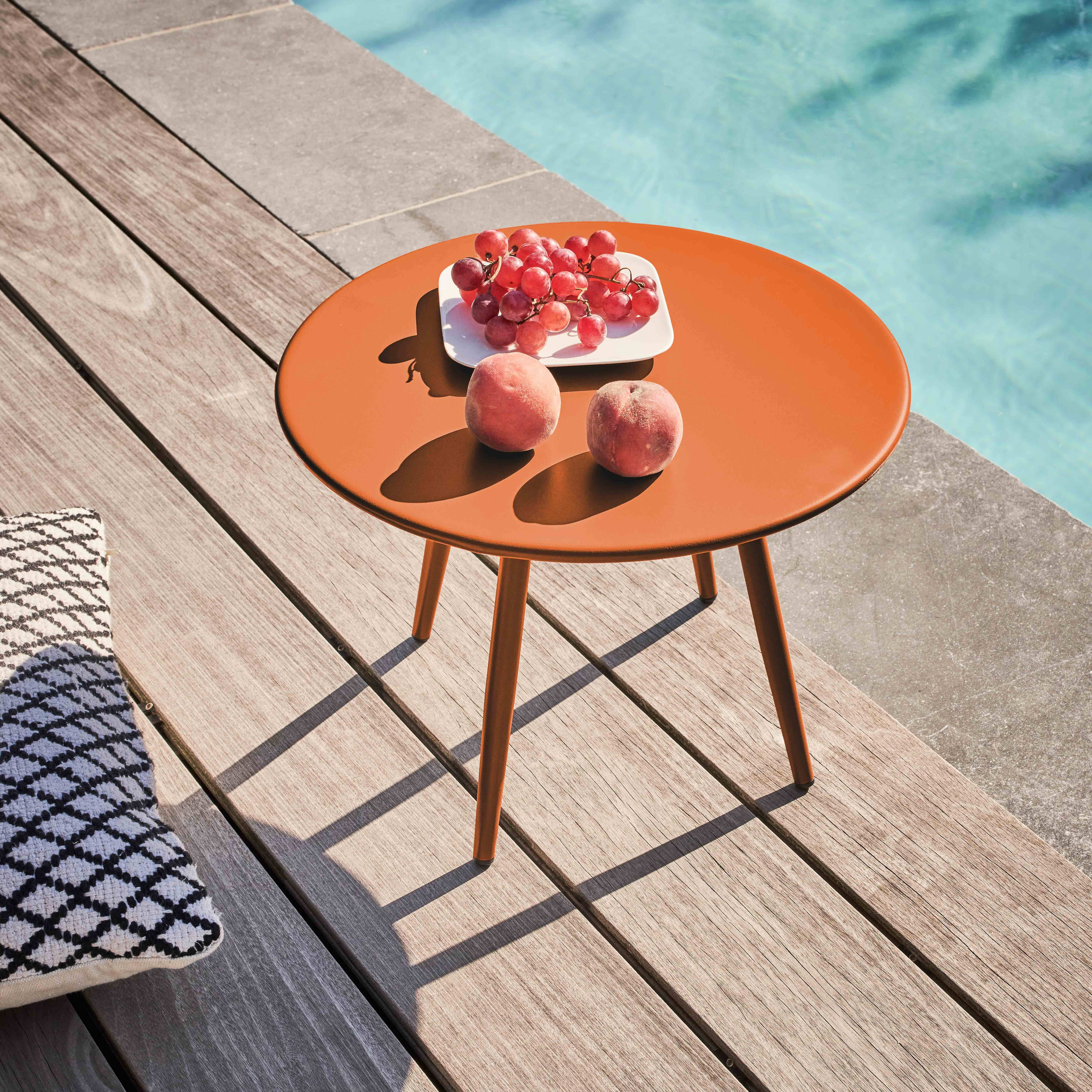 Ein orangefarbener Tisch mit Trauben und Pfirsichen auf einem Holzdeck, im Hintergrund ein Kissen und ein Pool.