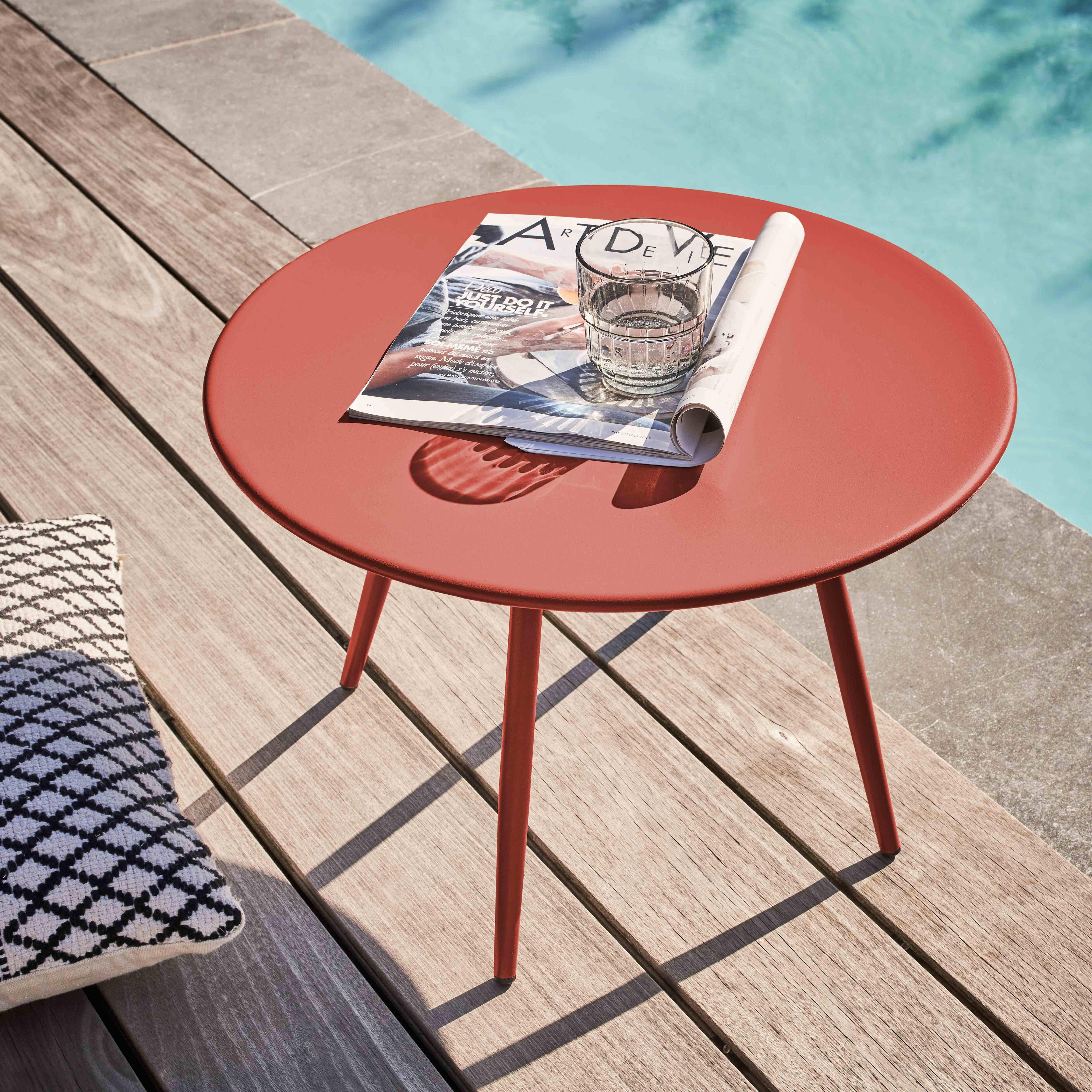 Ein roter Tisch mit einer Zeitschrift und einem Glas steht auf einem Deck in der Nähe eines Pools. Ein Kissen befindet sich links.