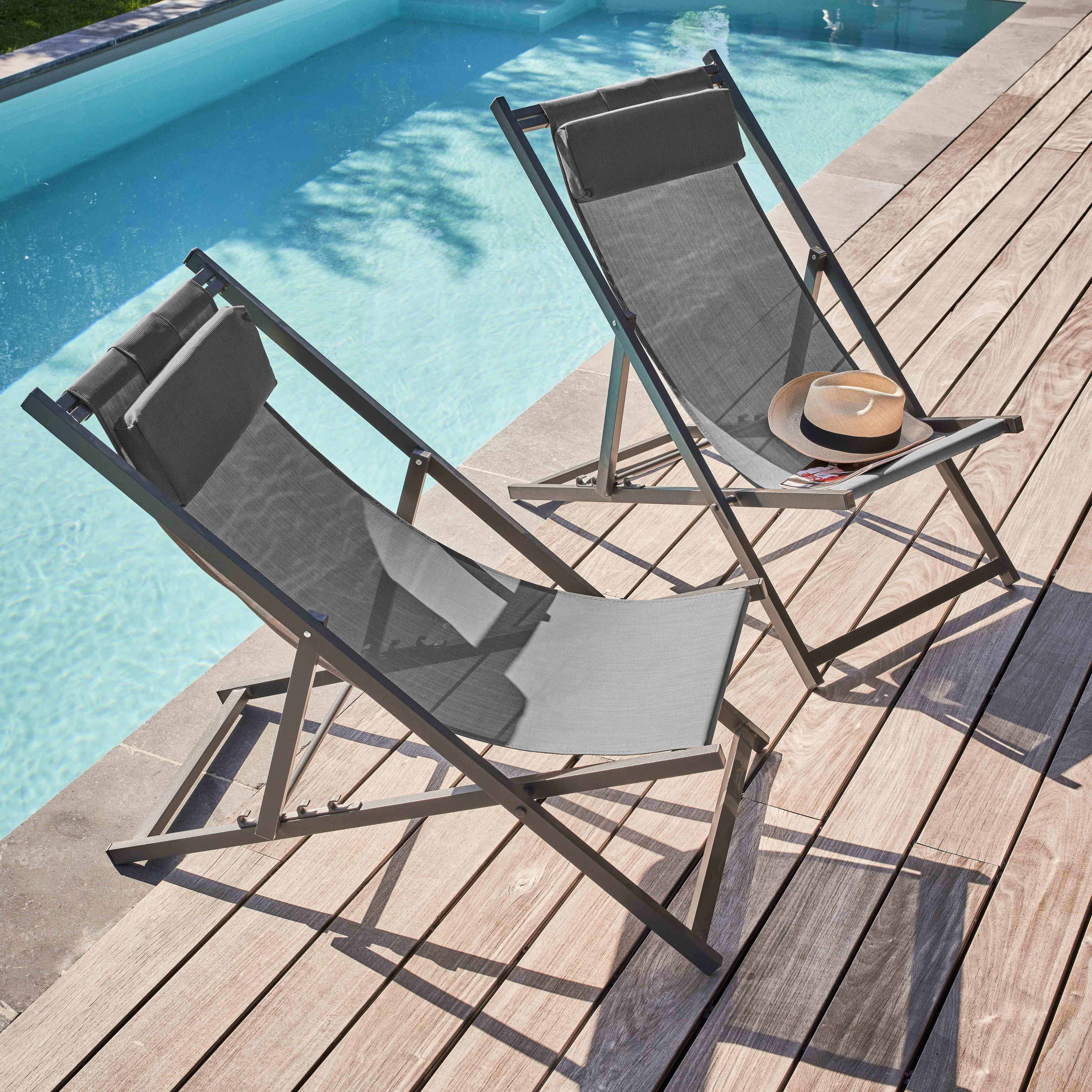 Zwei graue Liegestühle auf einem Holzdeck neben einem Swimmingpool. Auf einem Stuhl liegt ein Hut.