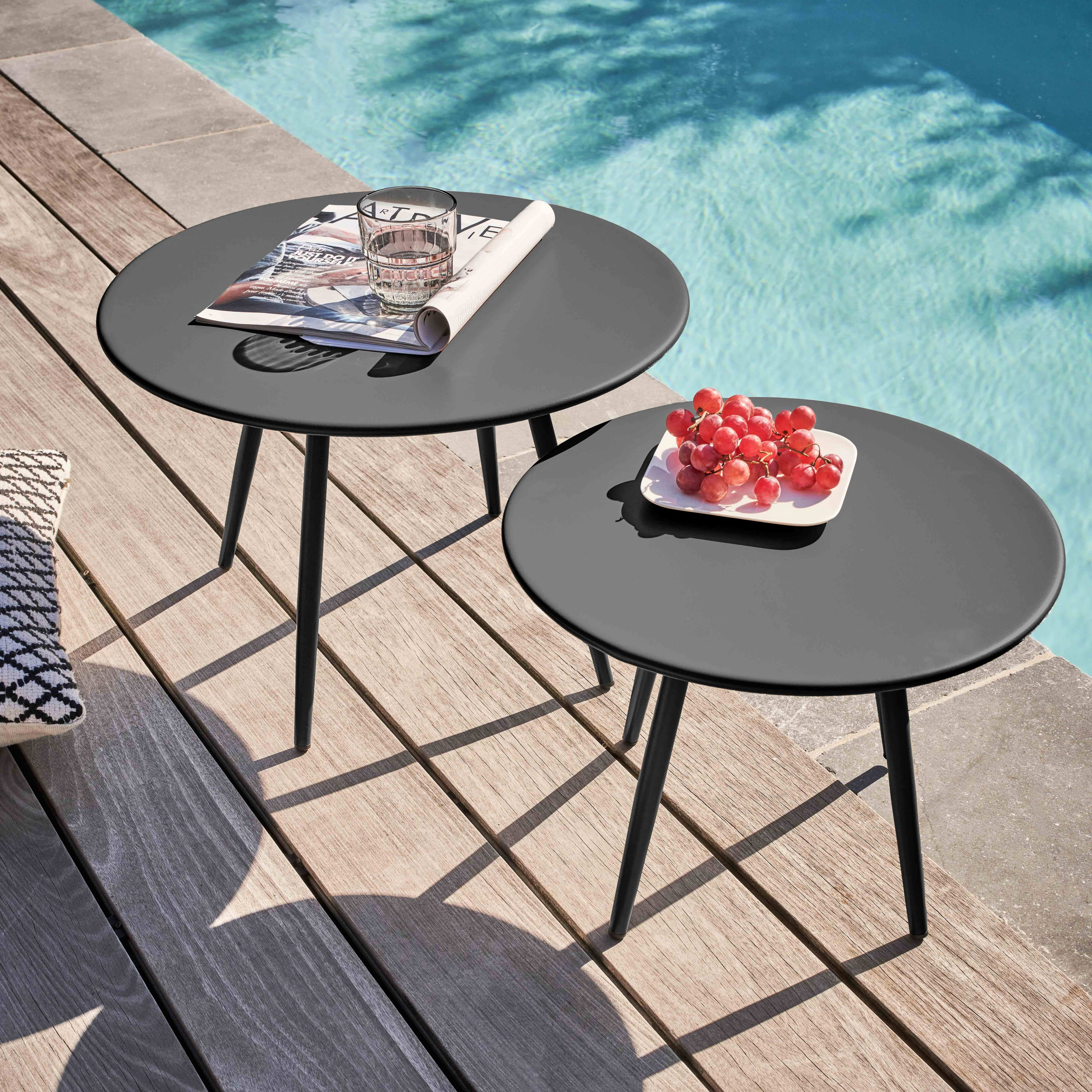Zwei schwarze Tische mit Gegenständen auf einem Holzdeck am Pool. Einer hat ein Glas und eine Zeitschrift, der andere Trauben.