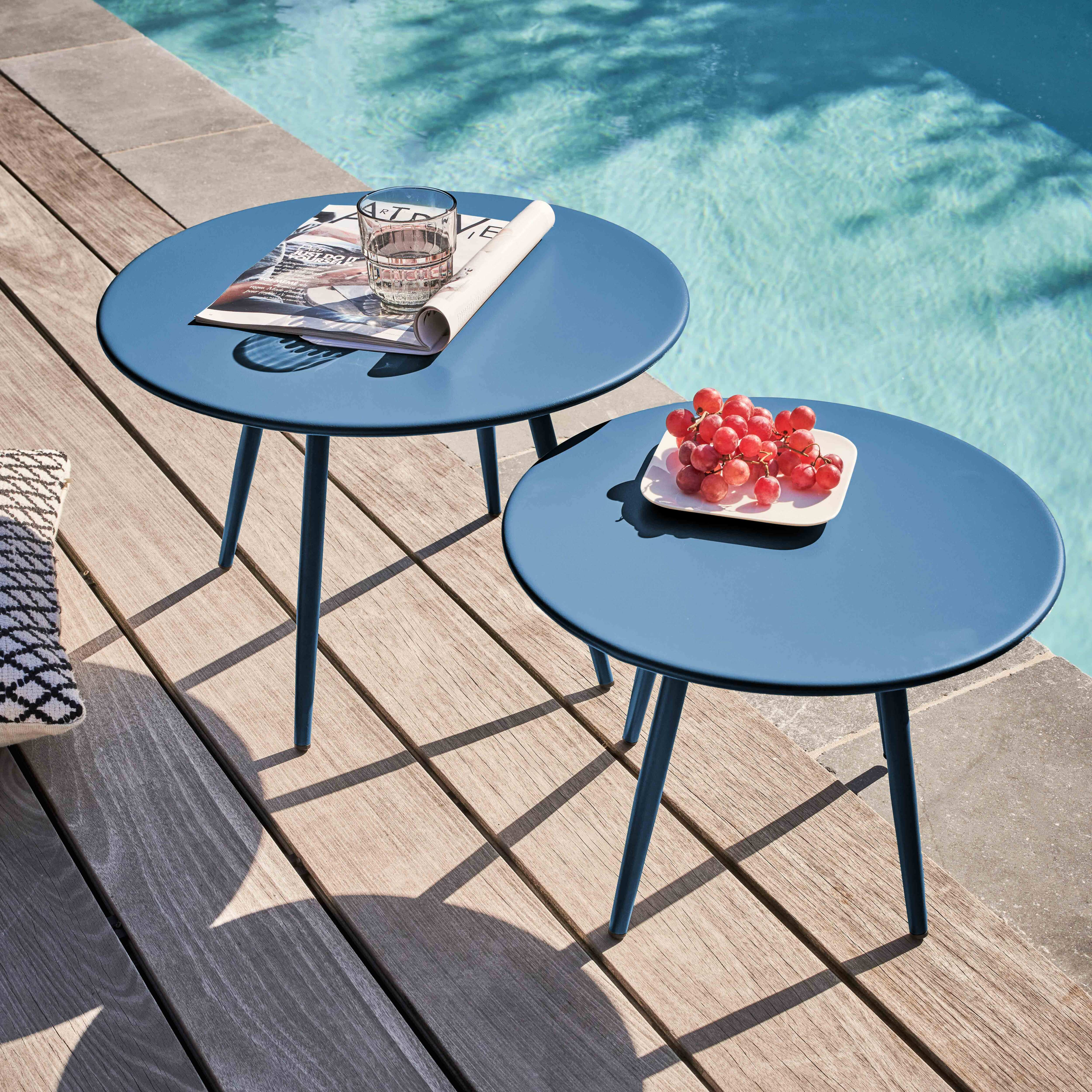 Zwei blaue Tische mit einer Zeitschrift und Trauben stehen an einem Pool. Holzterrasse umgibt den Pool. Schatten werden geworfen.
