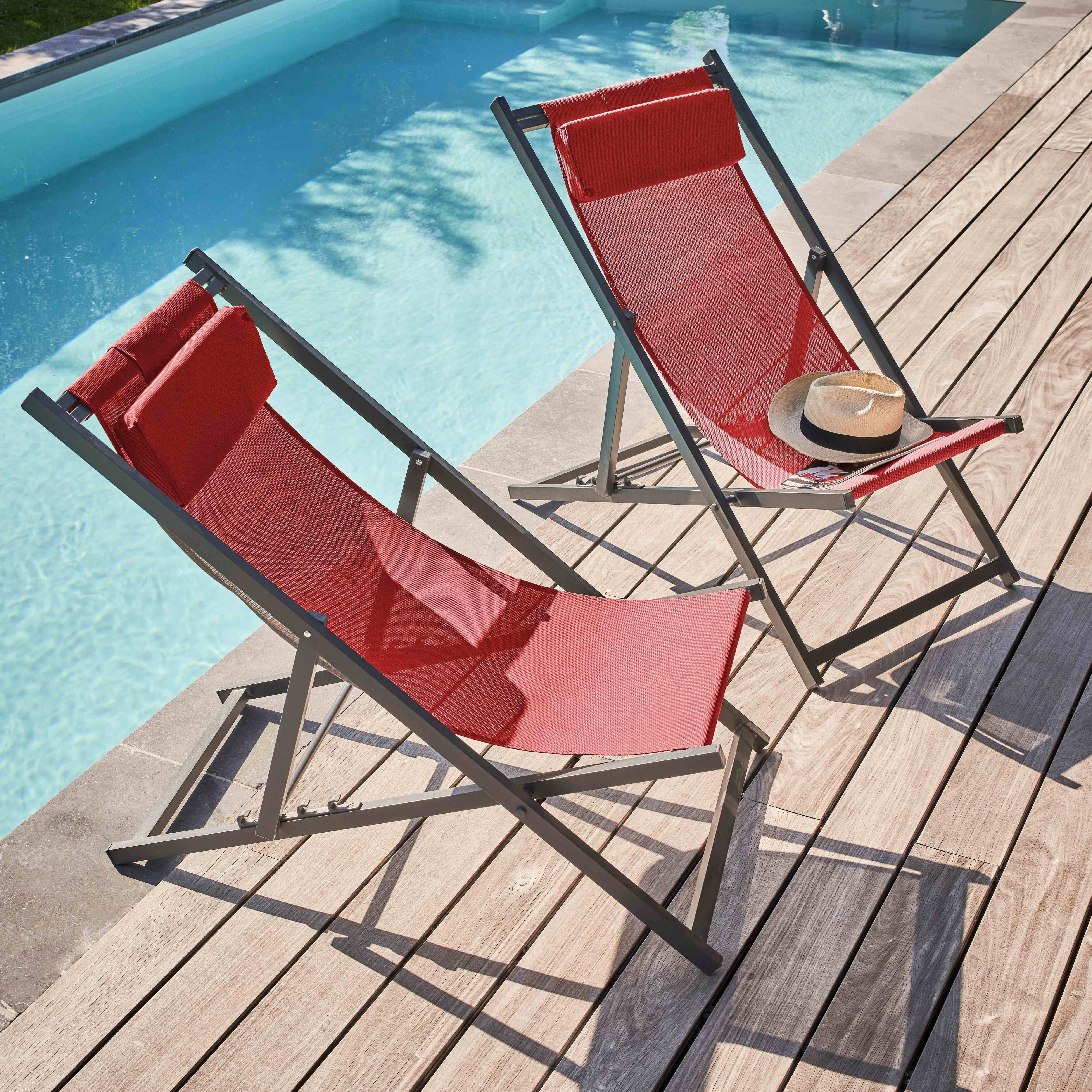 Zwei rote Liegestühle auf Holzterrasse, in der Nähe eines Pools. Auf einem Stuhl liegt ein Hut.