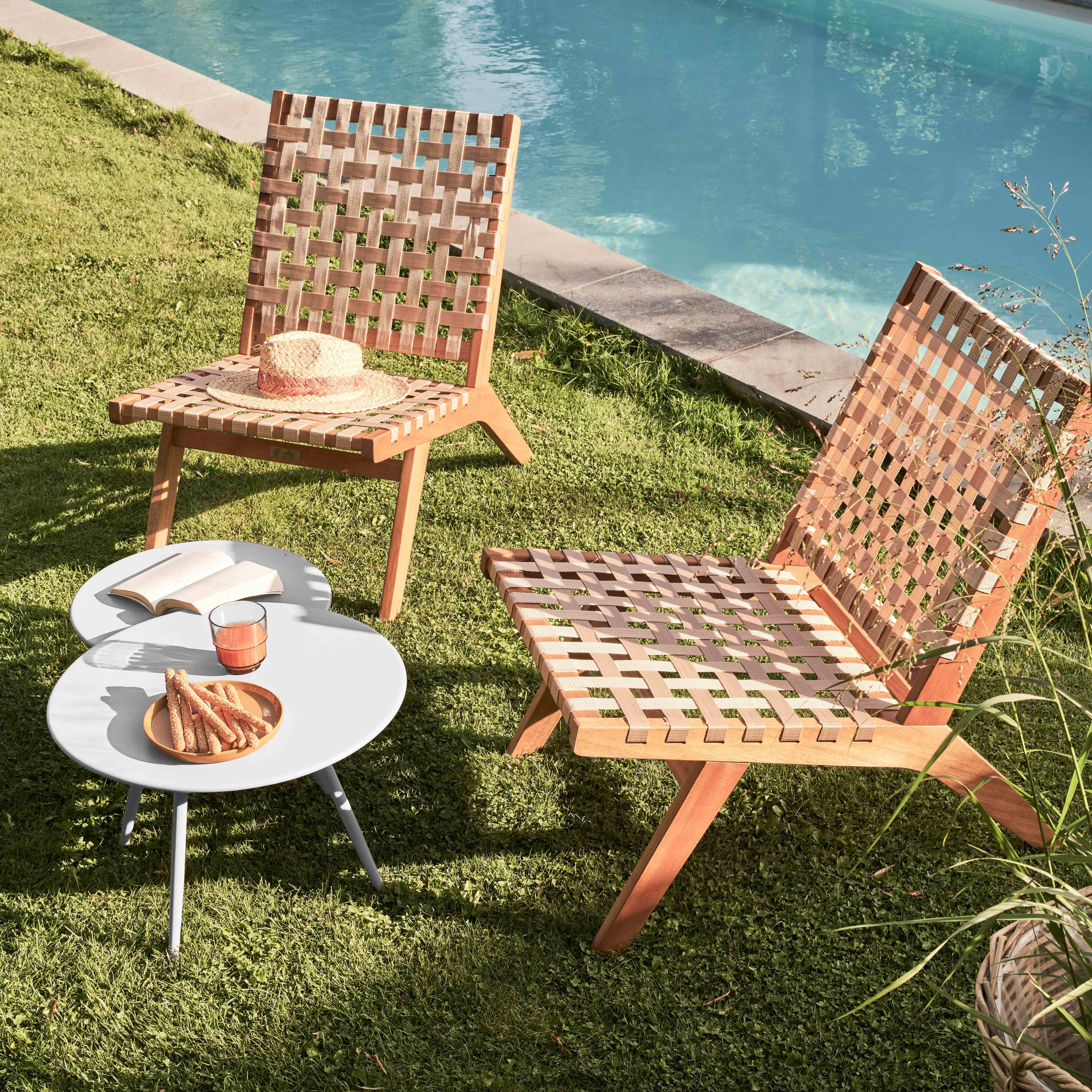 Zwei Holzstühle mit gewebten Sitzen auf Gras, in der Nähe eines Pools. Ein Tisch hat Snacks und Getränke. Ein Hut liegt auf einem Stuhl.