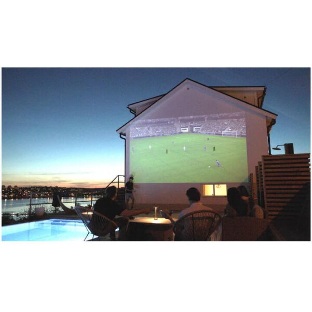 Leute sehen ein Fußballspiel, das auf eine Hauswand projiziert wird. Ein Pool, Stadtlichter und der Nachthimmel sind sichtbar.