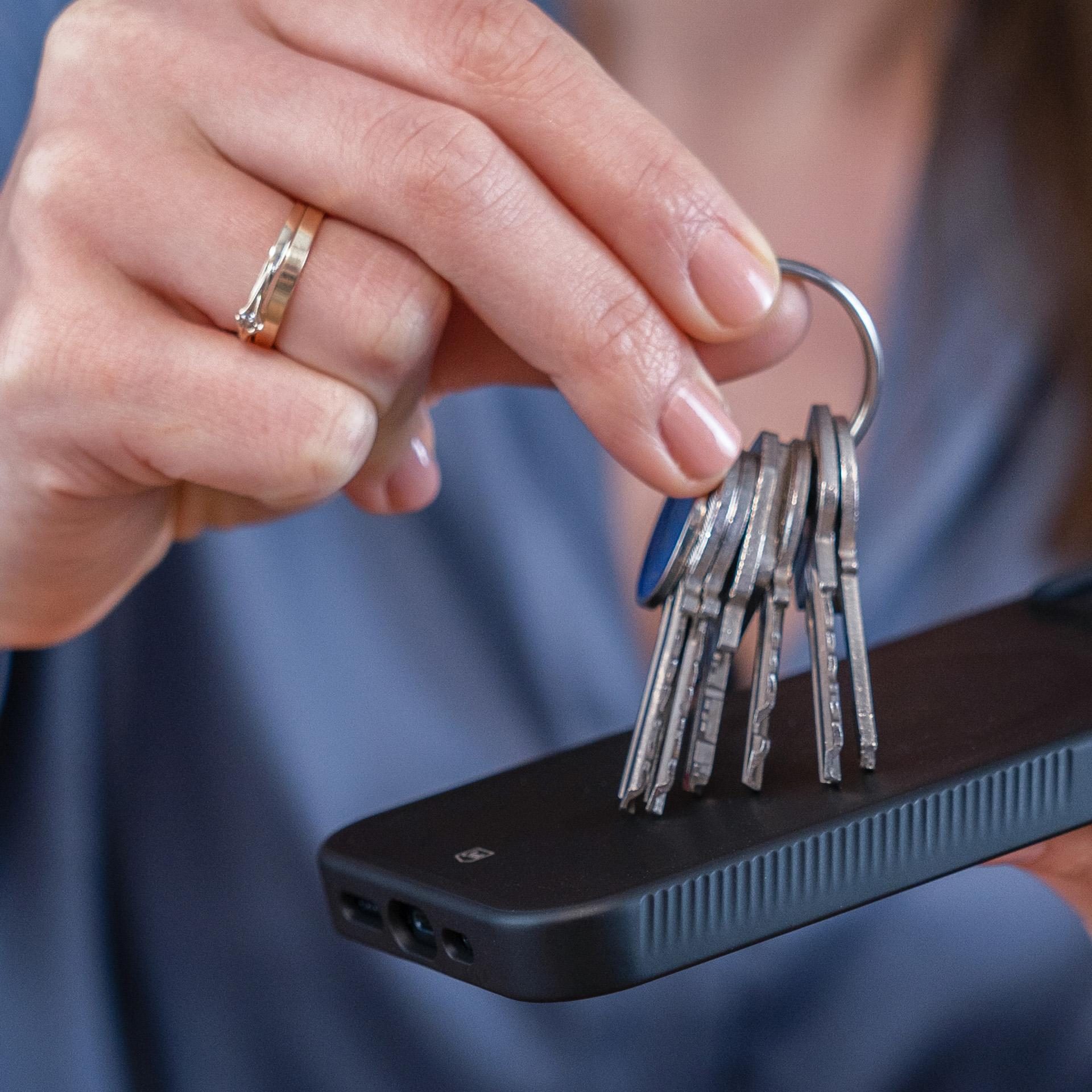 Ręka trzyma klucze i telefon. Złoty pierścionek jest na palcu. Telefon ma czarne etui.