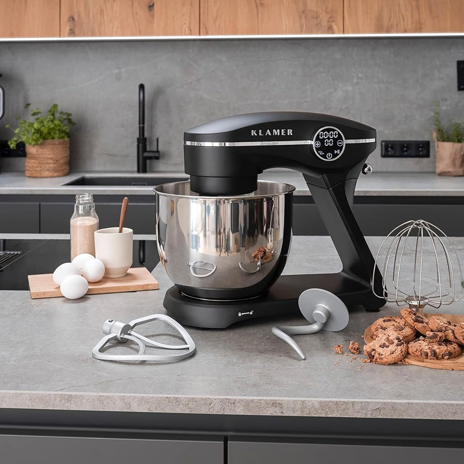 Ein schwarzer Küchenmixer mit Zubehör und Keksen auf einer grauen Arbeitsplatte. Eier, Mehl und Pflanzen sind im Hintergrund.