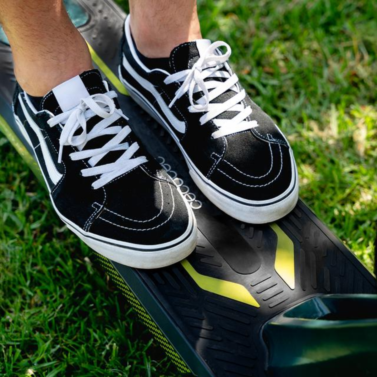 Zapatillas negras con cordones y rayas blancas sobre una patineta eléctrica negra y amarilla, sobre hierba verde.