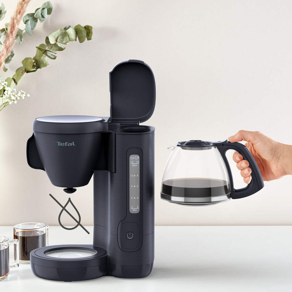 Bir elin tuttuğu cam bir sürahi ile siyah bir Tefal kahve makinesi. Ön planda iki fincan kahve.
