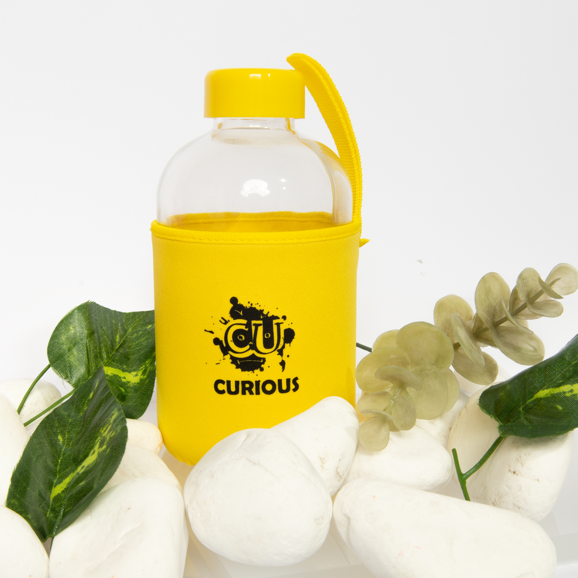 "Curious" yazılı sarı bir kılıf içinde bir su şişesi, beyaz taşların üzerinde, yeşil yapraklar ve bitkilerle.