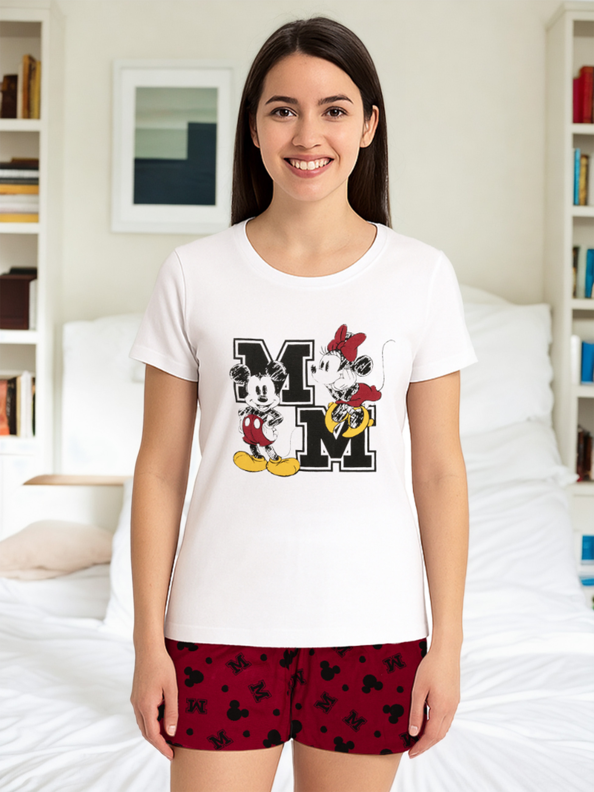 Frau in weißem T-Shirt mit Mickey- und Minnie-Mouse-Aufdruck, trägt rote Shorts. Hintergrund: Schlafzimmer.