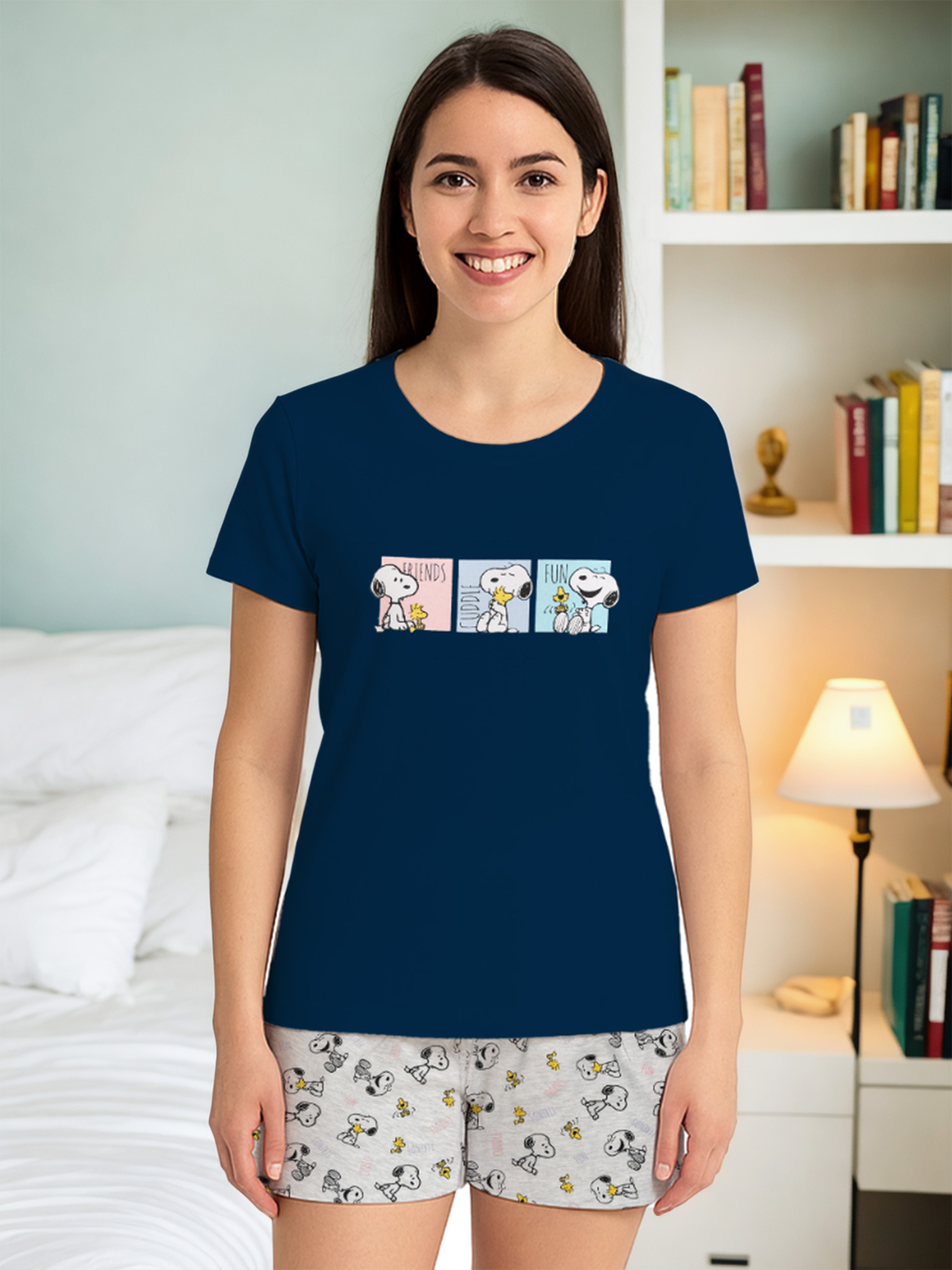 Eine Frau mit einem dunkelblauen Snoopy-T-Shirt und Shorts lächelt. Hinter ihr sind ein Bücherregal und ein Bett.