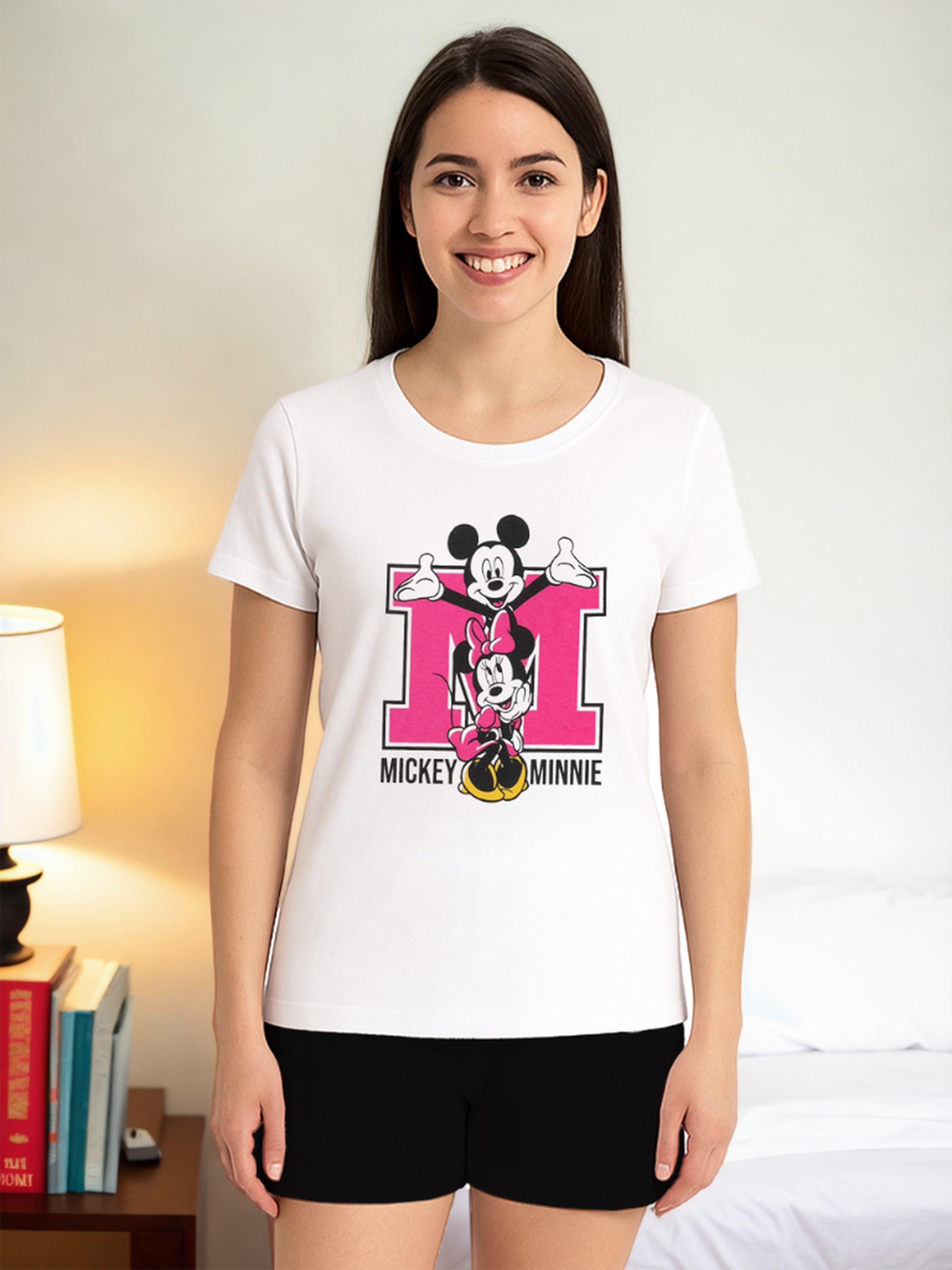 Frau mit weißem T-Shirt mit Mickey und Minnie Mouse. Sie steht in einem Zimmer mit Bett und Lampe.