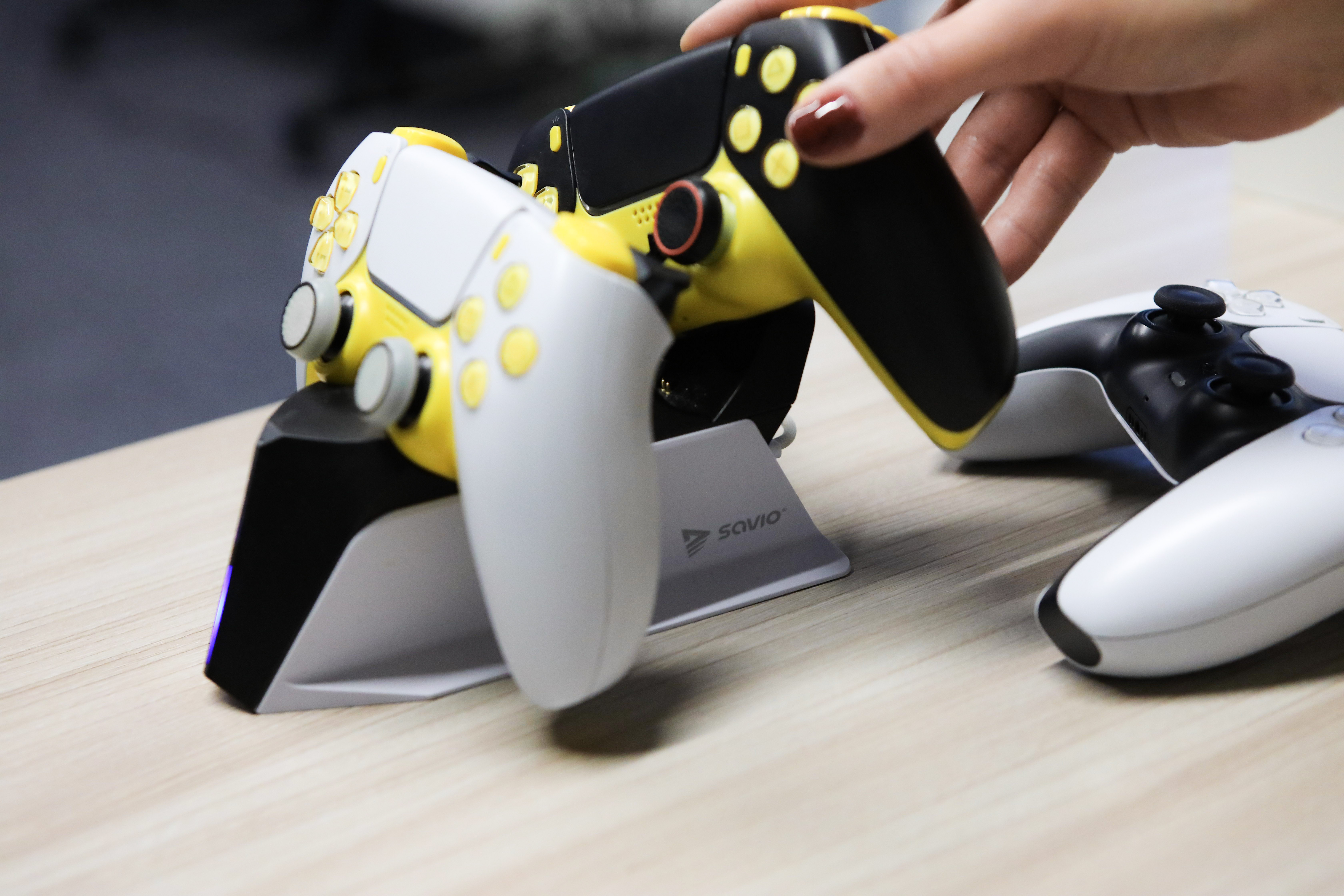 Dłoń trzyma czarno-żółty gamepad. Drugi biały gamepad na drewnianym stole i stacja ładująca.