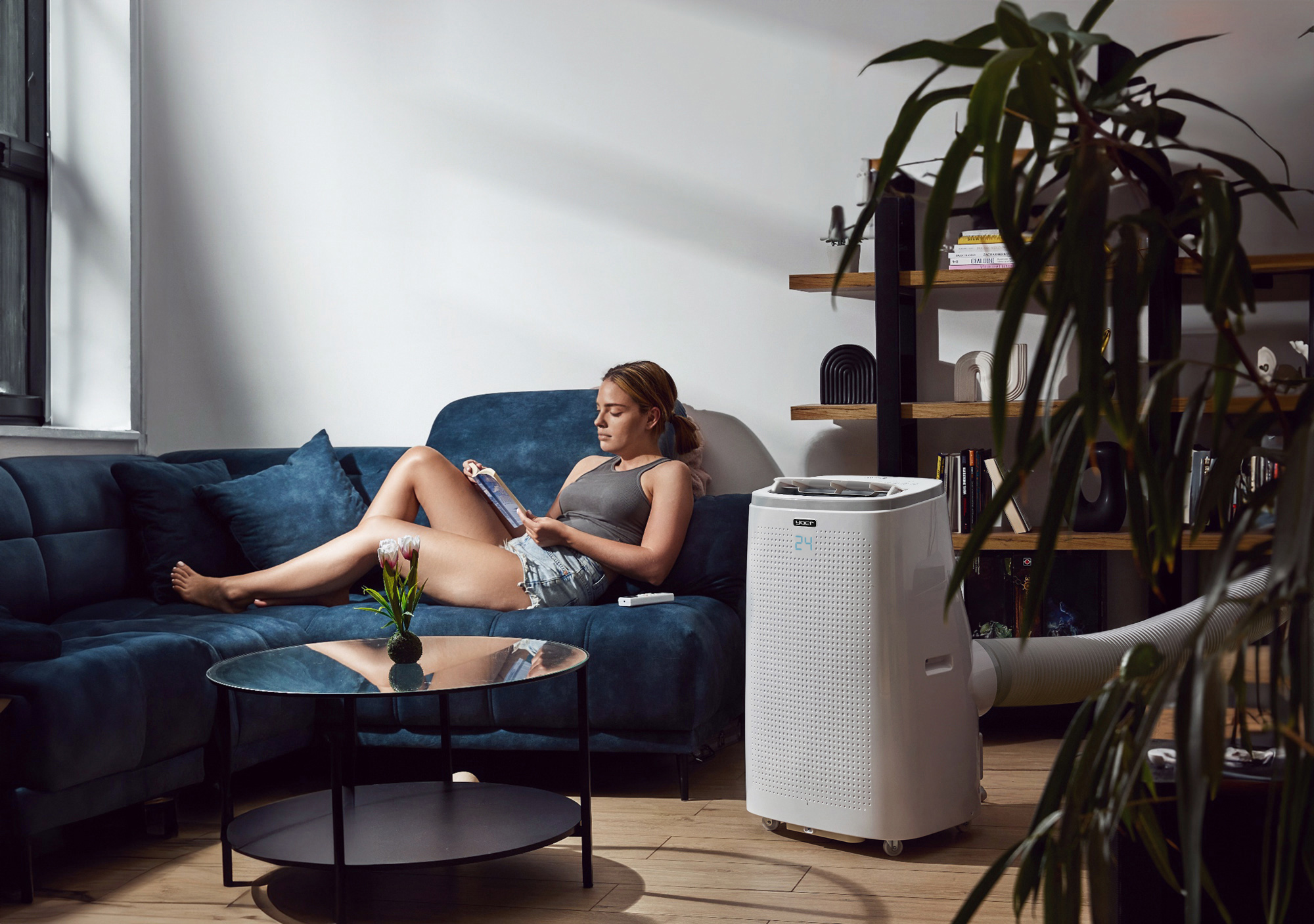 Kobieta na niebieskiej sofie czyta, obok klimatyzator. Stolik kawowy z kwiatami, półka na książki i okno w tle.